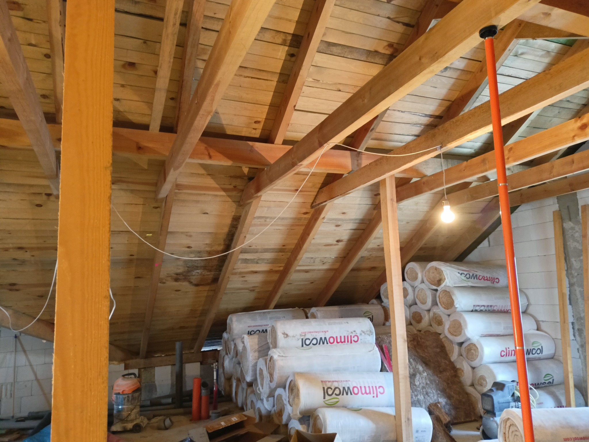 Poddasze w trakcie ocieplania, widoczne drewniane belki konstrukcyjne, stosy wełny mineralnej Climowool w foliowych opakowaniach, pomarańczowa rura podporowa i lampa wisząca na kablu.