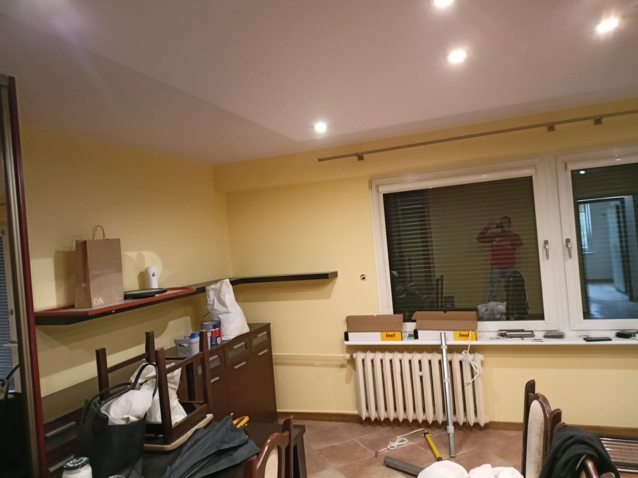 Pokój w trakcie przygotowań do prac wykończeniowych, widoczne materiały budowlane, narzędzia, meble przesunięte na środek, żółte ściany i osoba robiąca zdjęcie widoczna w odbiciu szyby okiennej.