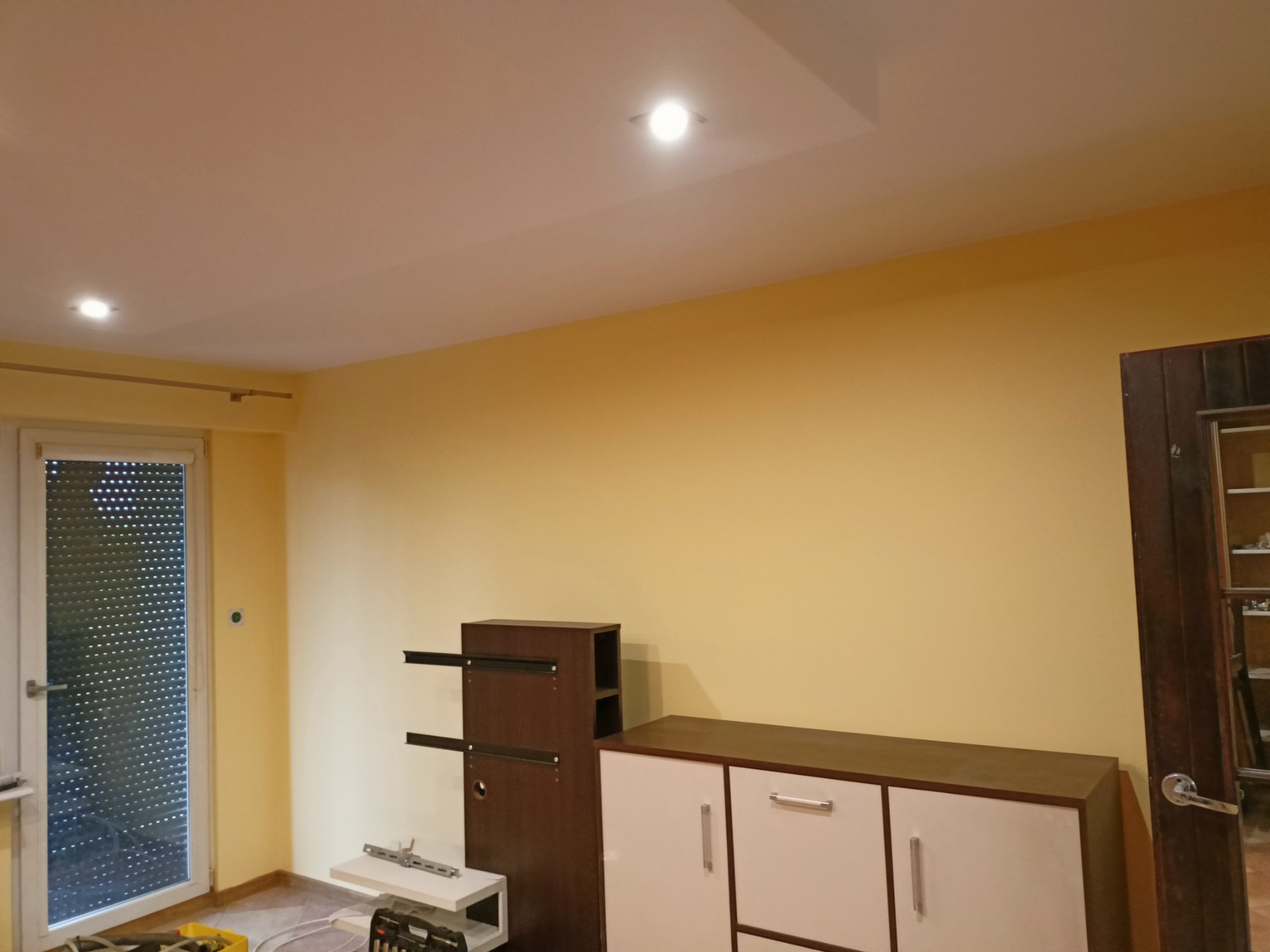 Żółta ściana w pokoju z zamontowanymi szafkami, widoczne narzędzia i akcesoria do montażu mebli, okno z roletami i sufit z oświetleniem punktowym.