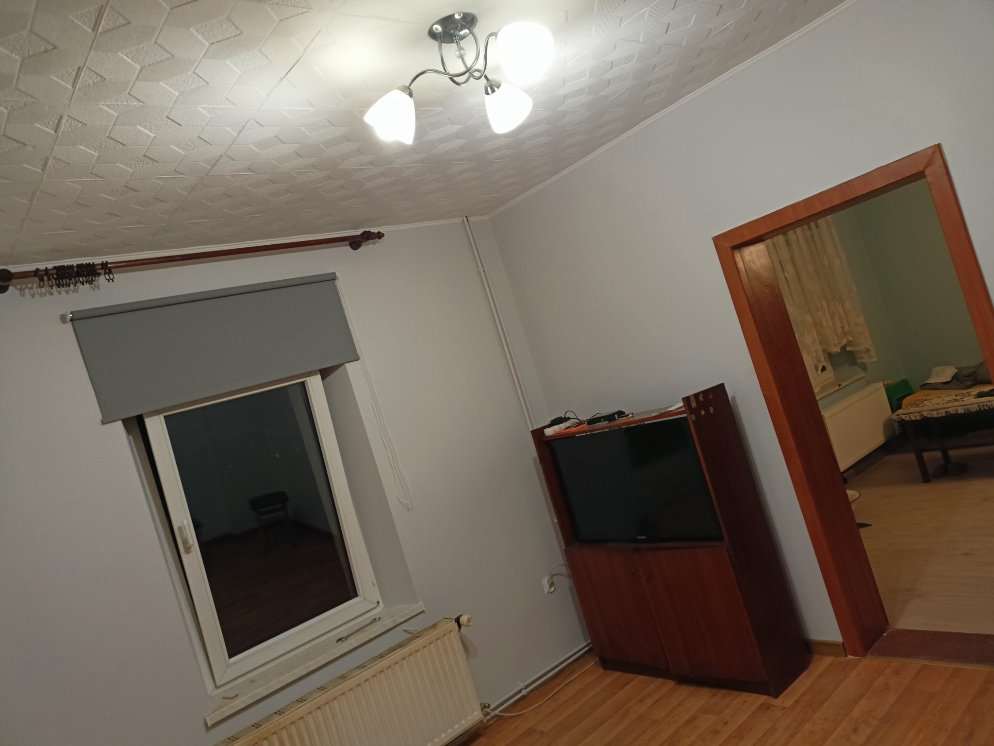 Pokój z drewnianą podłogą, grzejnikiem pod oknem z szarą roletą, telewizorem na szafce i widokiem na inne pomieszczenie przez otwarte drzwi z drewnianą ościeżnicą. Na suficie widoczna biała...