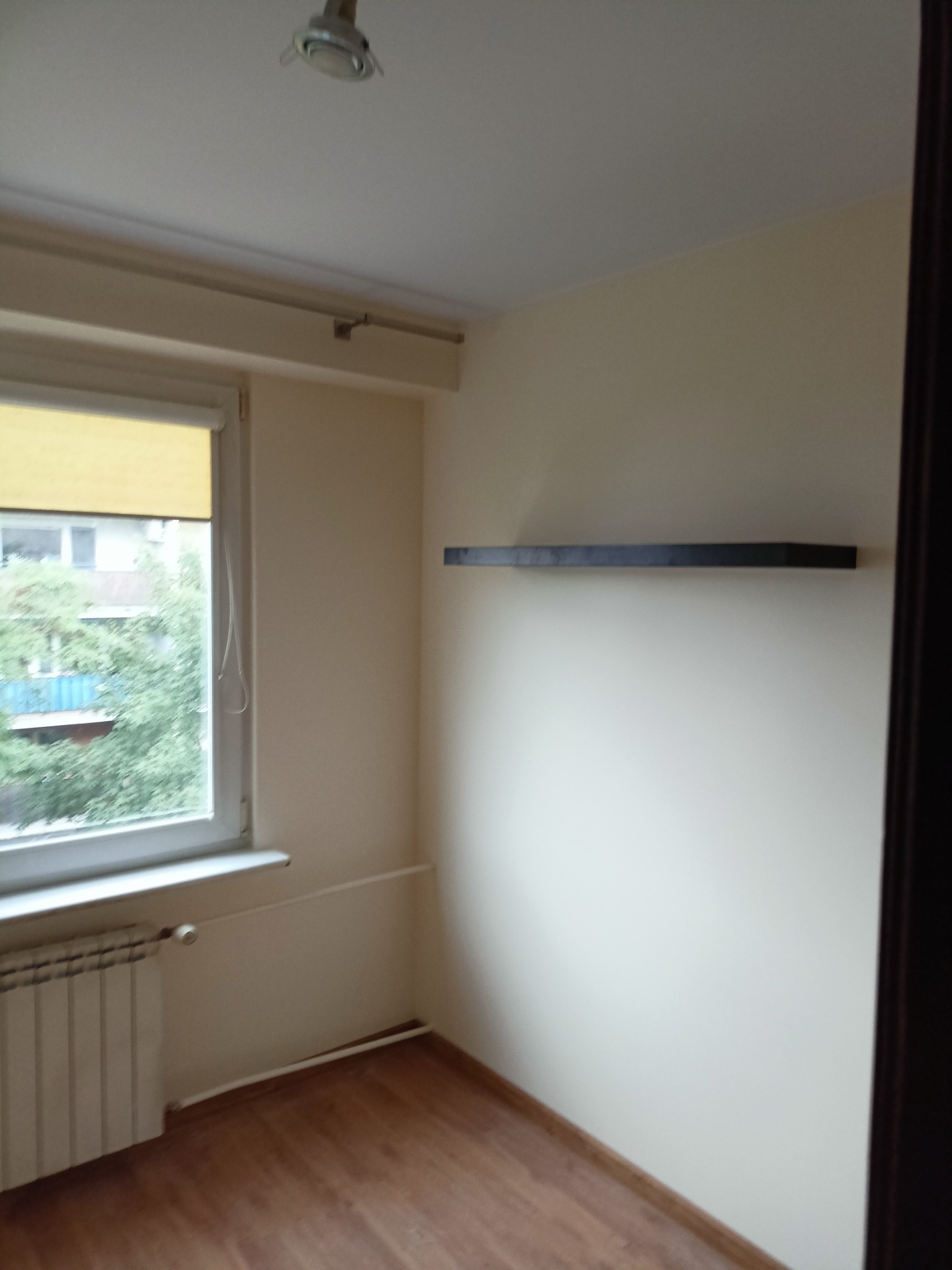 Pusty pokój po odświeżeniu: panele podłogowe, pomalowane ściany na jasny kolor, grzejnik pod oknem z żółtą roletą, pojedyncza czarna półka na ścianie.