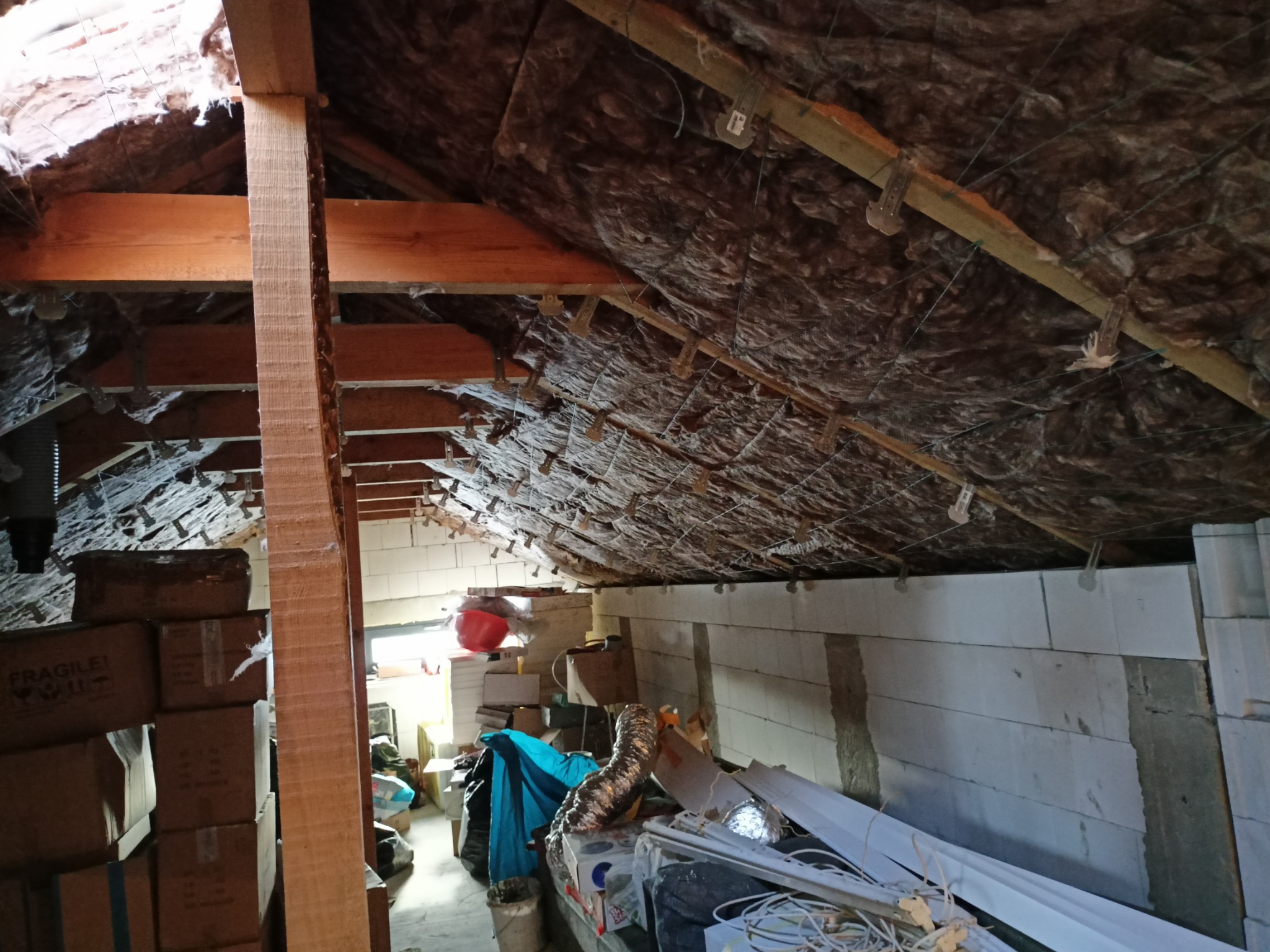 Poddasze ocieplone brązową wełną mineralną, widoczne drewniane belki konstrukcyjne, białe ściany z bloczków oraz kartony i różne przedmioty składowane w przestrzeni.