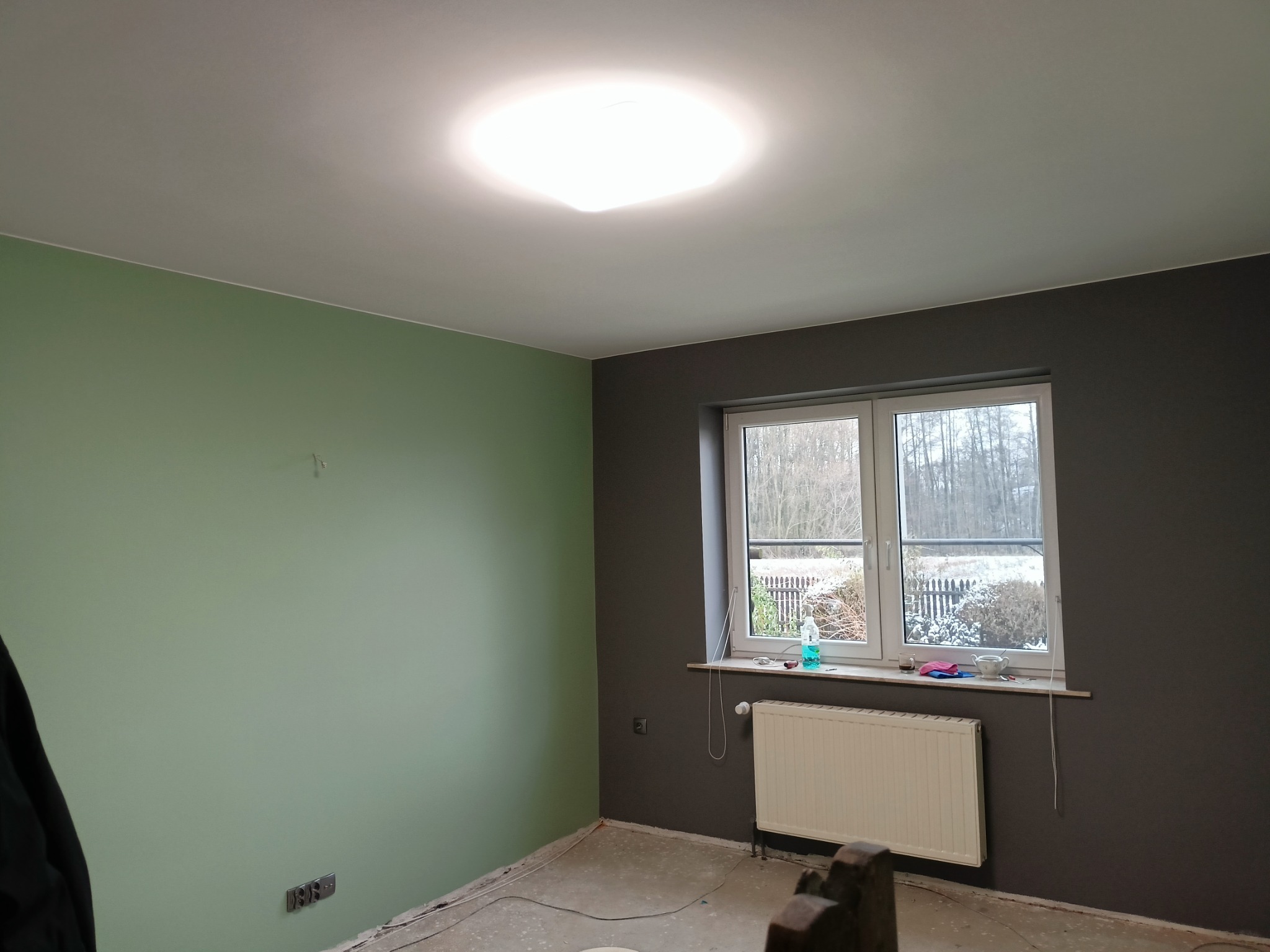 Pokój po malowaniu z jedną ścianą zieloną, drugą grafitową, z oknem i grzejnikiem, widoczne ślady po remoncie na podłodze.