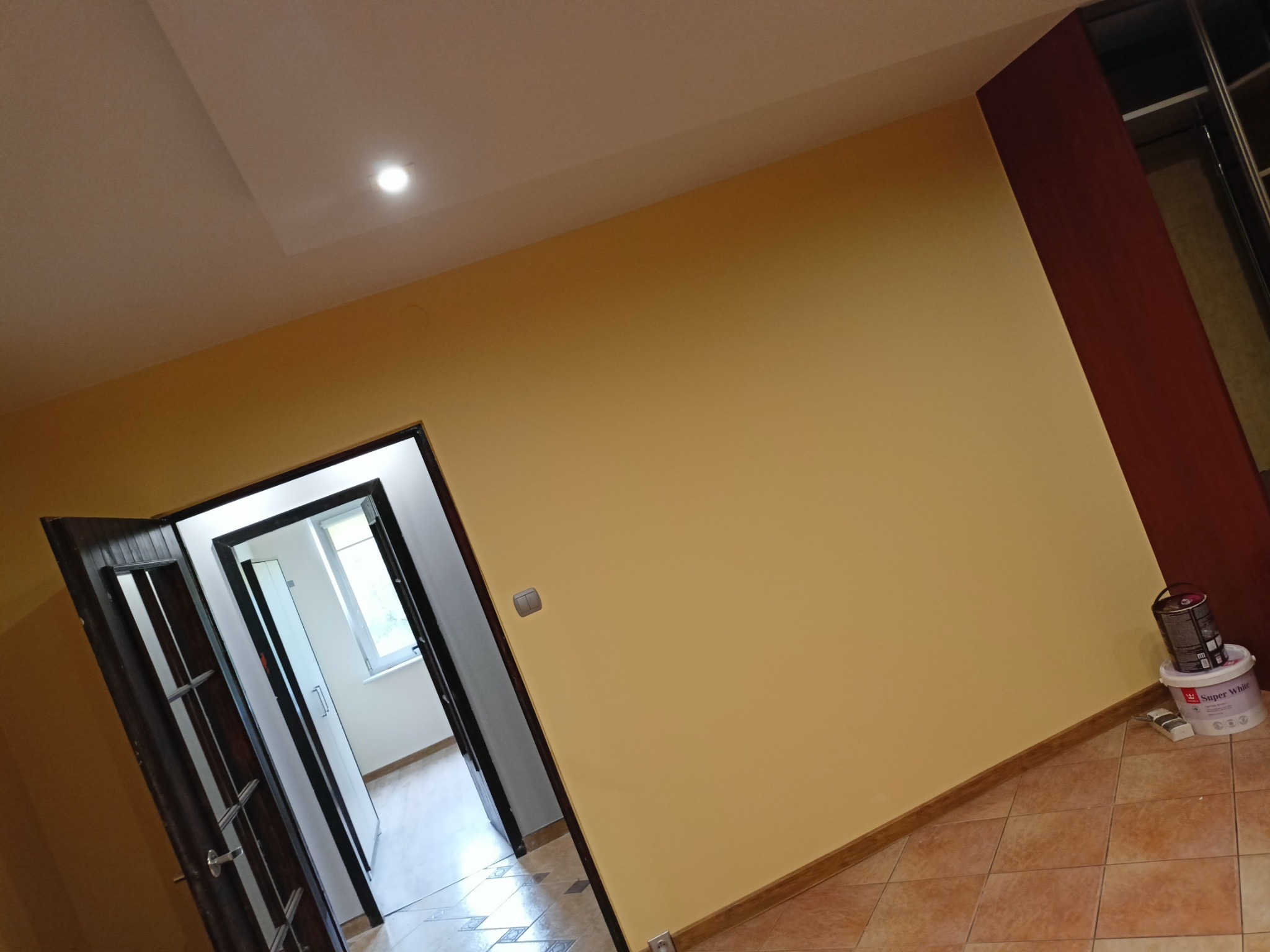 Pomalowane na żółto wnętrze z otwartymi drzwiami, widoczna puszka z farbą Super White przy ścianie, brązowe płytki podłogowe i drewniana szafa w rogu.