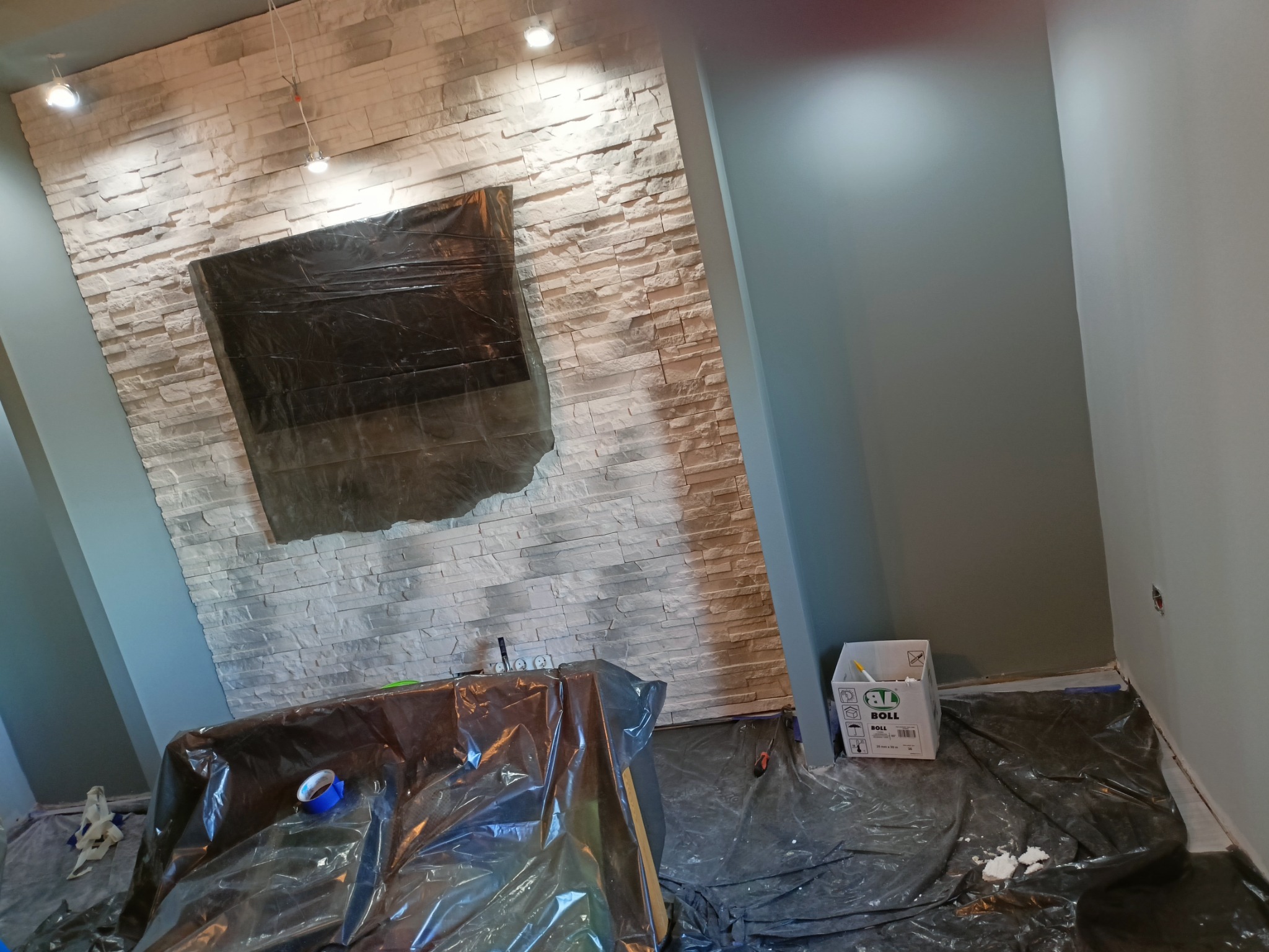 Pokój w trakcie przygotowań do malowania: ściana z dekoracyjną okładziną kamienną zabezpieczona folią, podłoga również zakryta folią ochronną, widoczny karton z akcesoriami malarskimi marki Boll.