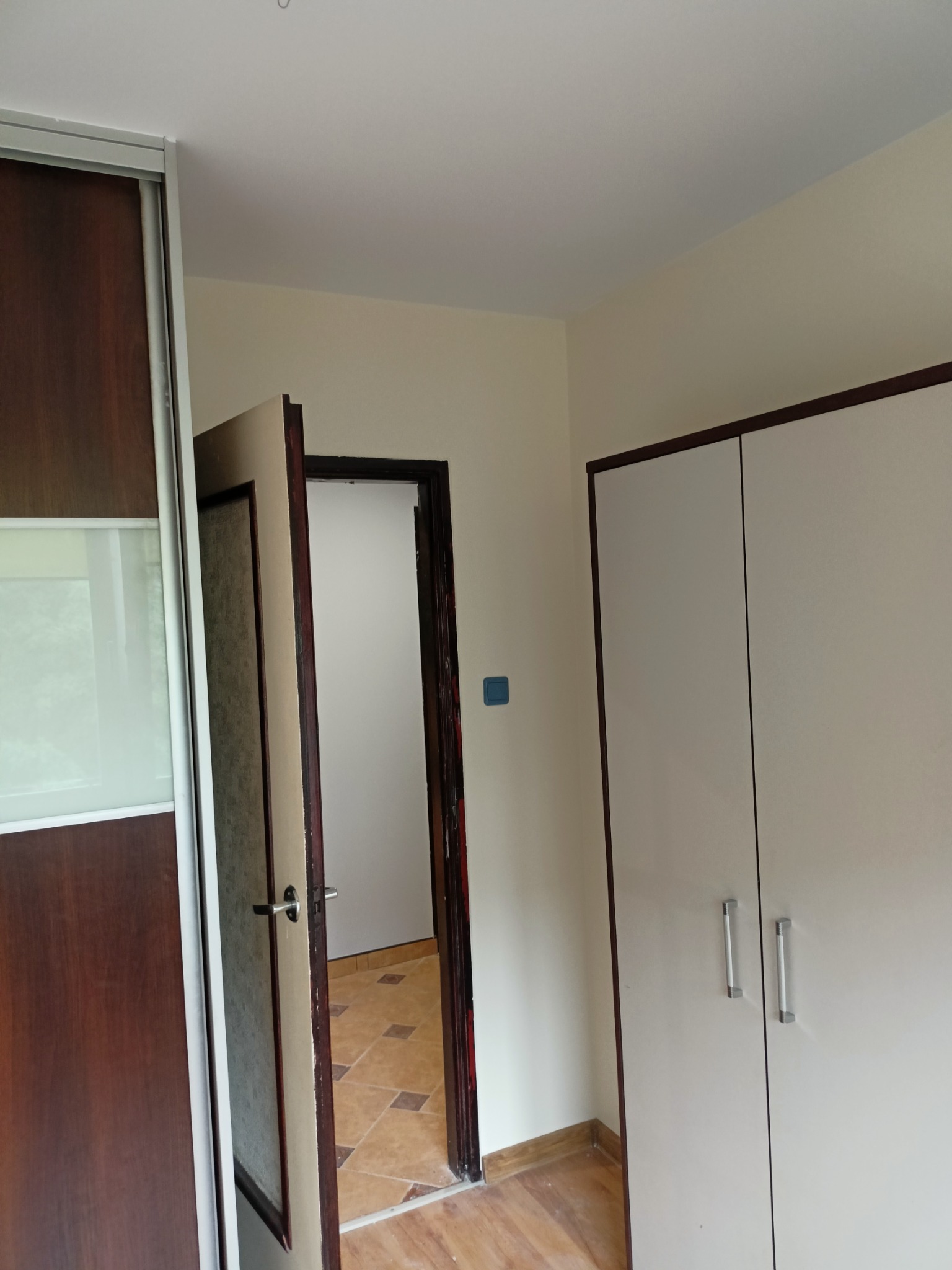 Wnętrze pokoju z widokiem na otwarte drzwi do kolejnego pomieszczenia z wzorzystą podłogą, po lewej szafa przesuwna z drewnianymi drzwiami i lustrem, po prawej biała szafa z dwoma drzwiami.