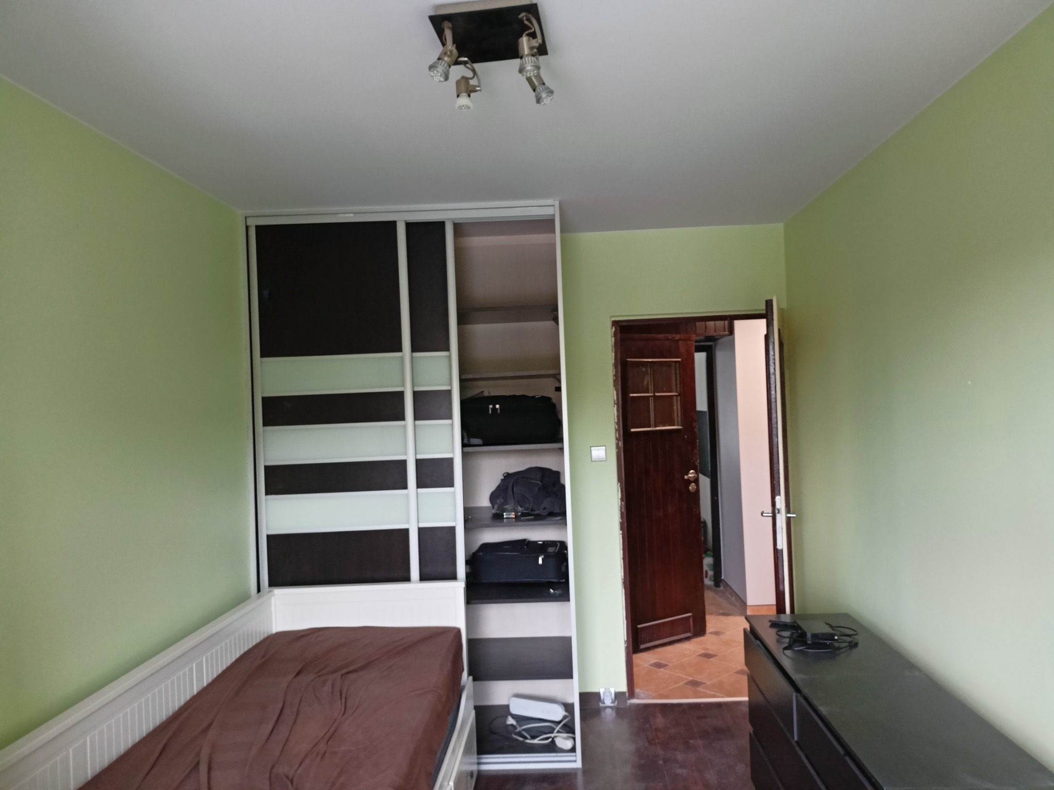 Pokój z łóżkiem z brązową pościelą, szafą z przesuwnymi drzwiami, komodą i otwartymi drzwiami do innego pomieszczenia, ściany pomalowane na jasnozielono.