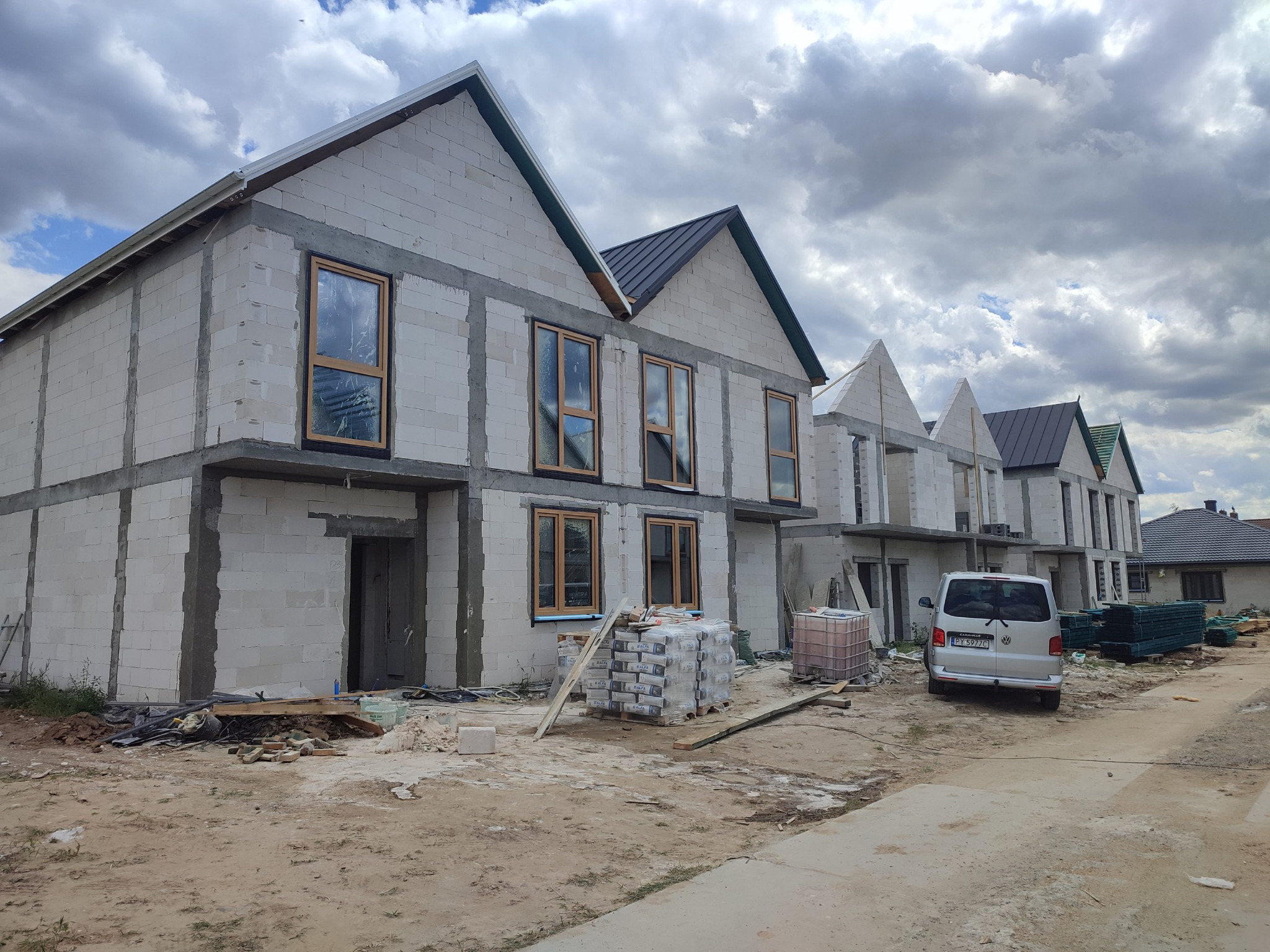 Surowy stan budowy szeregu domów z betonu komórkowego, widoczne okna i materiały budowlane. Częściowo wykończone dachy, na posesji zaparkowany bus.