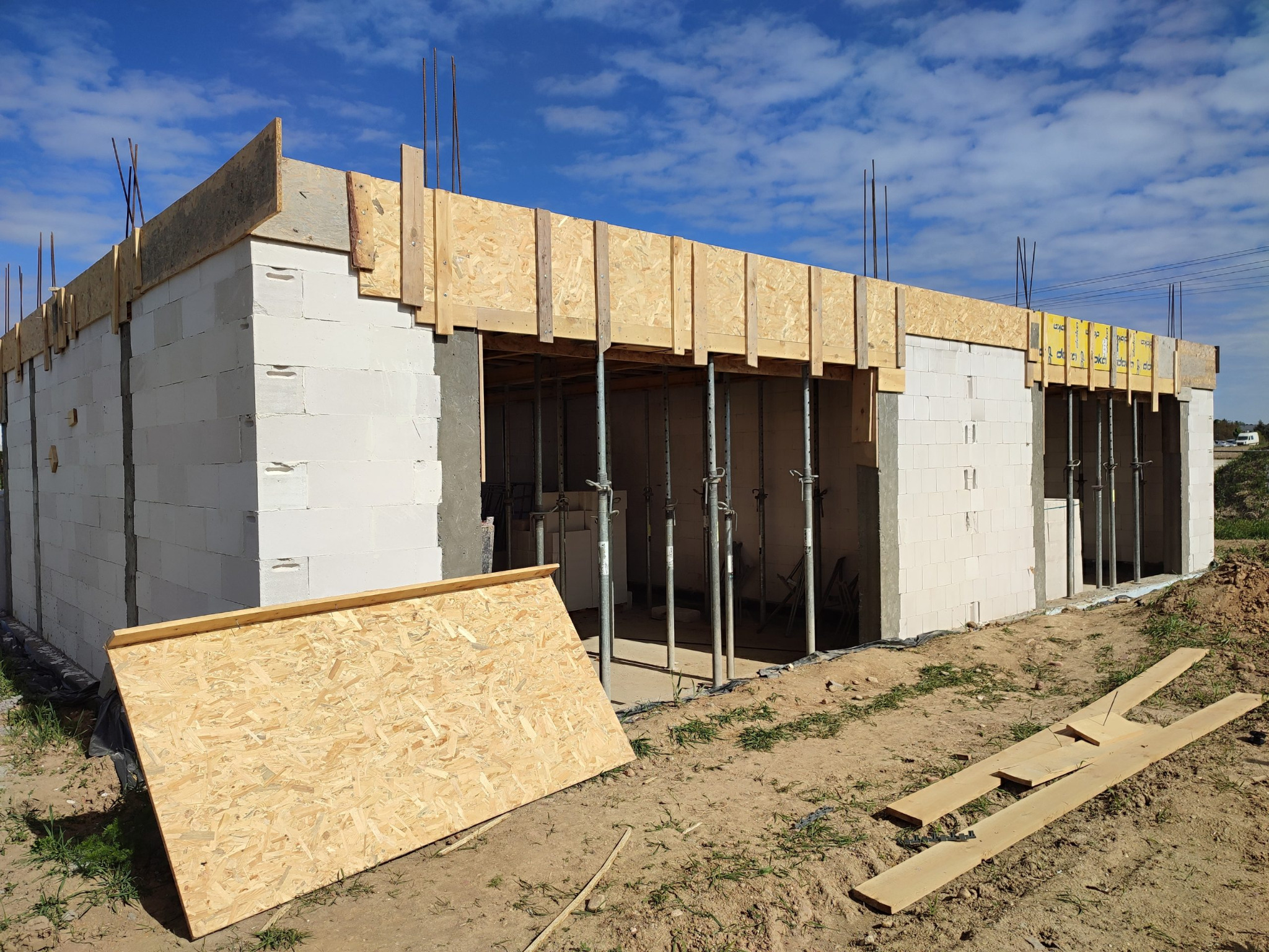 Wznoszenie ścian z betonu komórkowego. Widoczne szalunki i podpory stropu. Elementy drewniane na ziemi. Budowa w toku, słoneczny dzień, błękitne niebo z chmurami.