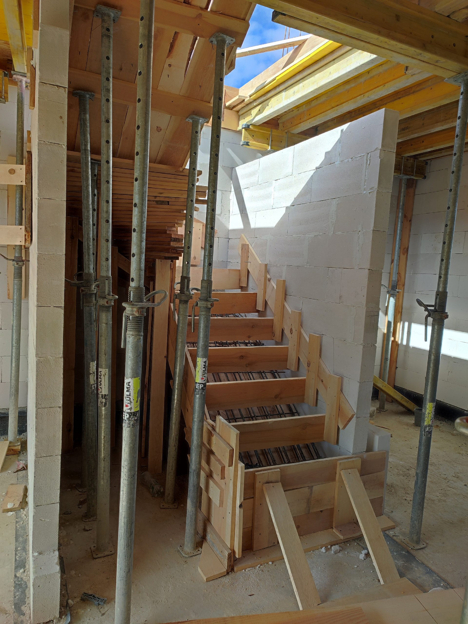 Wewnętrzne schody w trakcie budowy, zbrojenie i drewniane szalunki, wsparte metalowymi stemplami ULMA, ściany z bloczków betonu komórkowego.