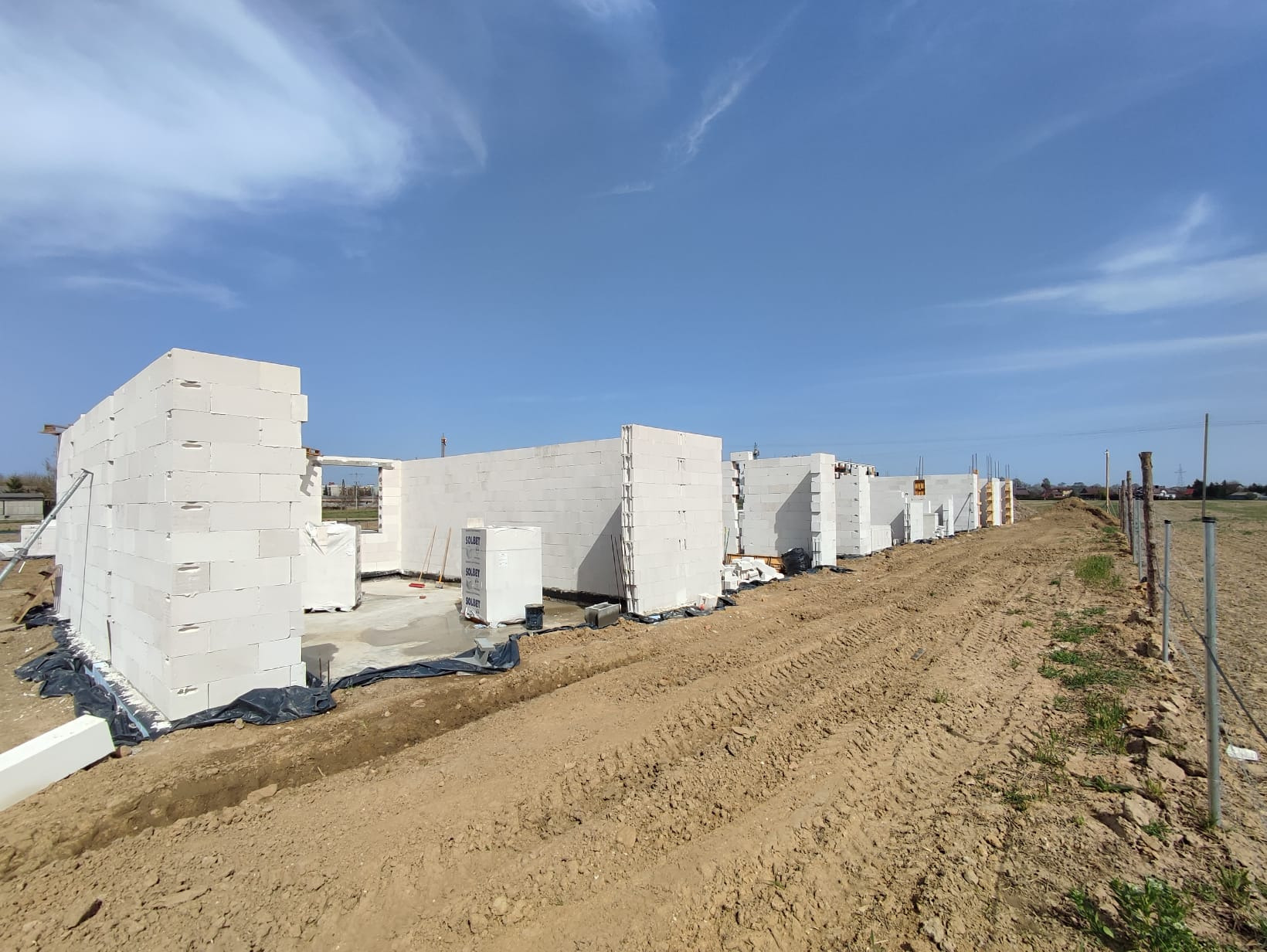 Budowa domu z białych bloczków Solbet, widok z zewnątrz na postęp prac, teren budowy z widocznymi śladami opon i błękitne niebo.