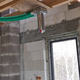 Instal-Grzew Grzegorz Salamon - Instalacja rekuperacji powietrza w surowym wnętrzu budynku, widoczne kanały wentylacyjne pod stropem i przewody elektryczne przy ścianie z oknem.