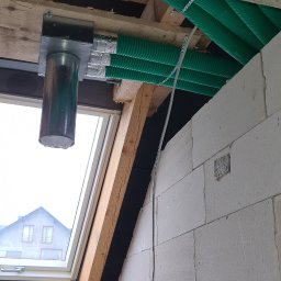 Instal-Grzew Grzegorz Salamon - Instalacja wentylacyjna z zielonymi, karbowanymi rurami poprowadzona pod stropem w surowym wnętrzu, widoczny fragment okna dachowego i ściana z białego betonu komórkowego.