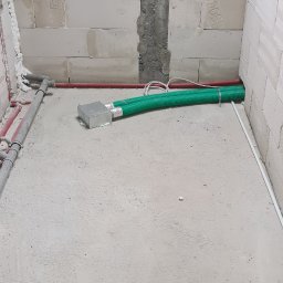 Instal-Grzew Grzegorz Salamon - Surowy stan pomieszczenia z widoczną instalacją ogrzewania podłogowego: zielone rury karbowane wychodzące ze srebrnej skrzynki, czerwone rury grzewcze wzdłuż ściany z bloczków betonowych, widoczne...