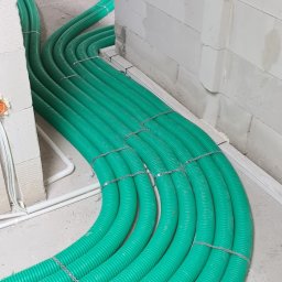 Instal-Grzew Grzegorz Salamon - Zielone, karbowane rury ogrzewania podłogowego, ułożone równolegle i przymocowane metalowymi paskami, biegnące wzdłuż surowej ściany z białych bloczków w trakcie instalacji.