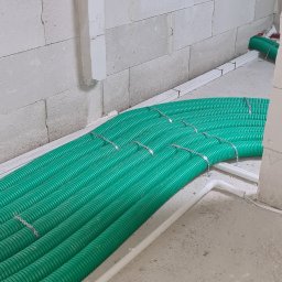 Instal-Grzew Grzegorz Salamon - Zielone rury instalacji grzewczej w trakcie montażu, przymocowane metalowymi uchwytami do podłoża w surowym stanie, ściany z białych bloczków.