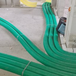 Instal-Grzew Grzegorz Salamon - Wiązka zielonych, karbowanych rur instalacyjnych ułożonych na surowym betonie w trakcie prac wykończeniowych w budynku z białych bloczków.