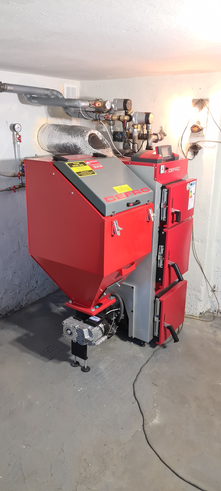 Czerwono-szary piec na paliwo stałe marki Defro z podajnikiem, widoczne rury instalacji grzewczej pod sufitem, w tle biała ściana i szara podłoga.