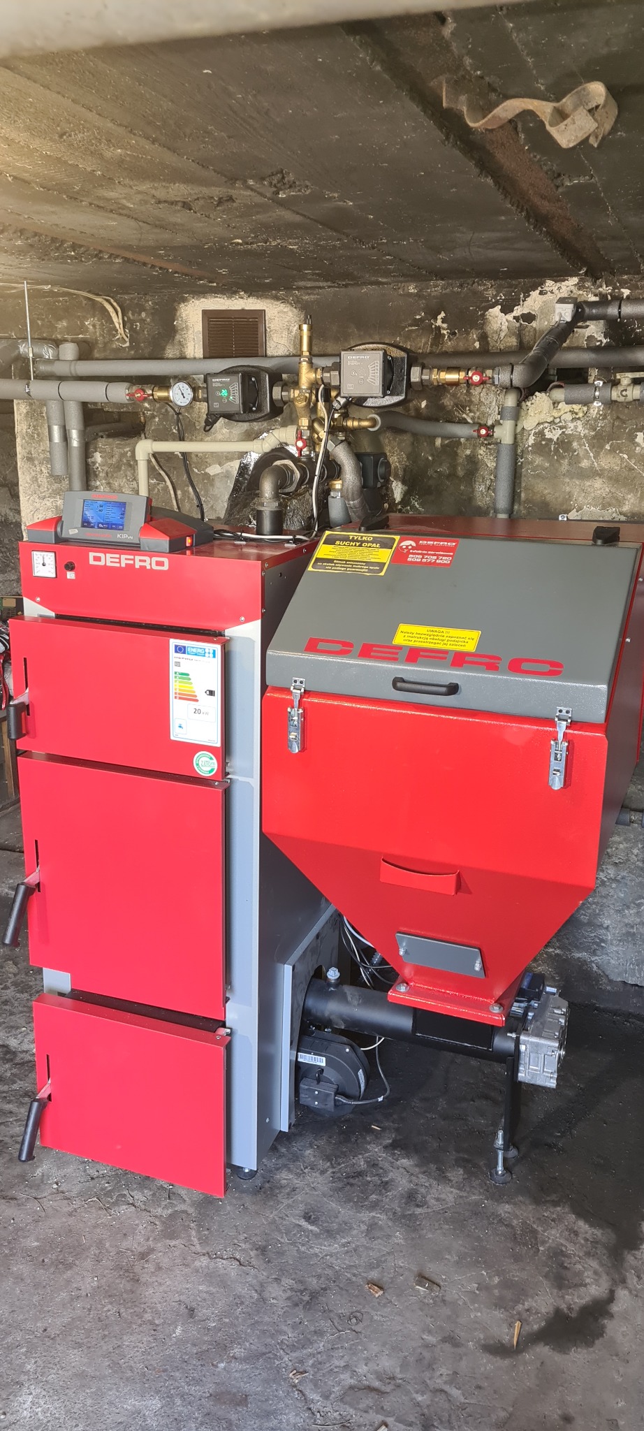 Czerwono-szary piec na paliwo stałe marki Defro z podajnikiem, zamontowany w piwnicy, widoczne rury instalacji grzewczej i sterownik.