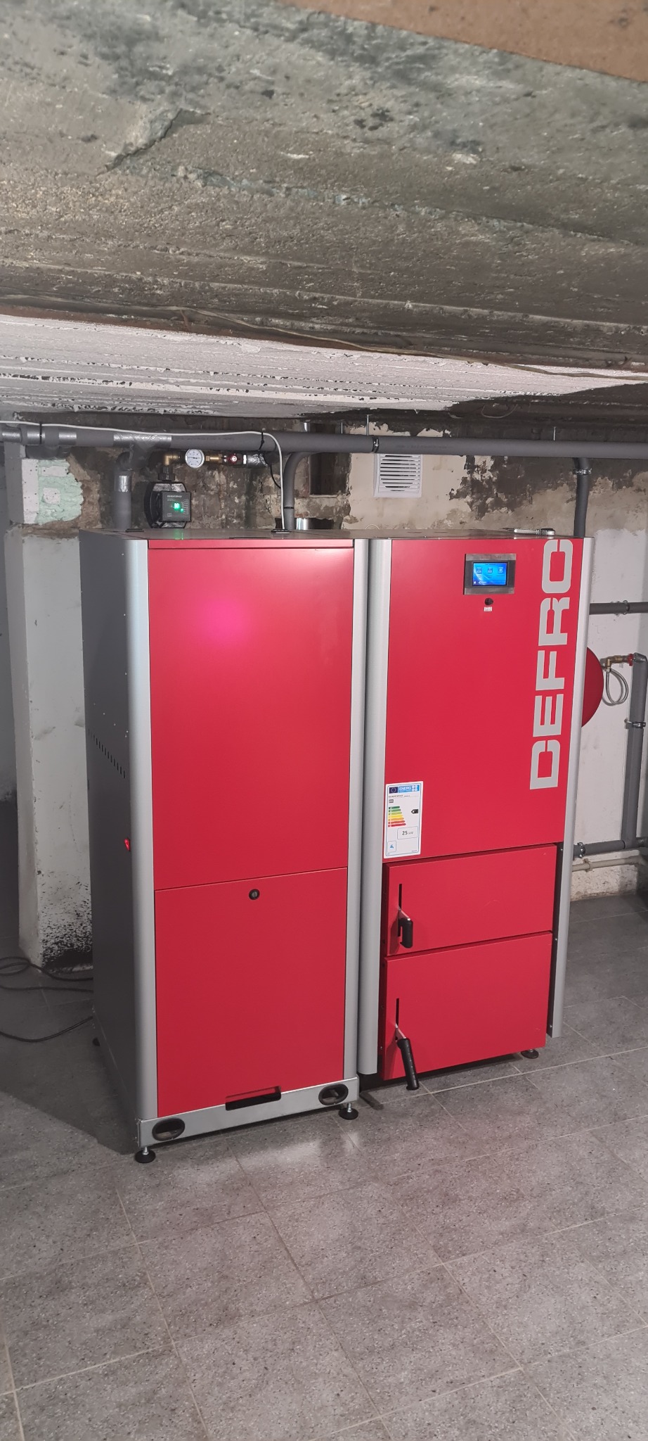 Czerwono-szary piec z podajnikiem na paliwo stałe marki DEFRO, zainstalowany w piwnicy z widoczną instalacją grzewczą na ścianie i betonowym sufitem.