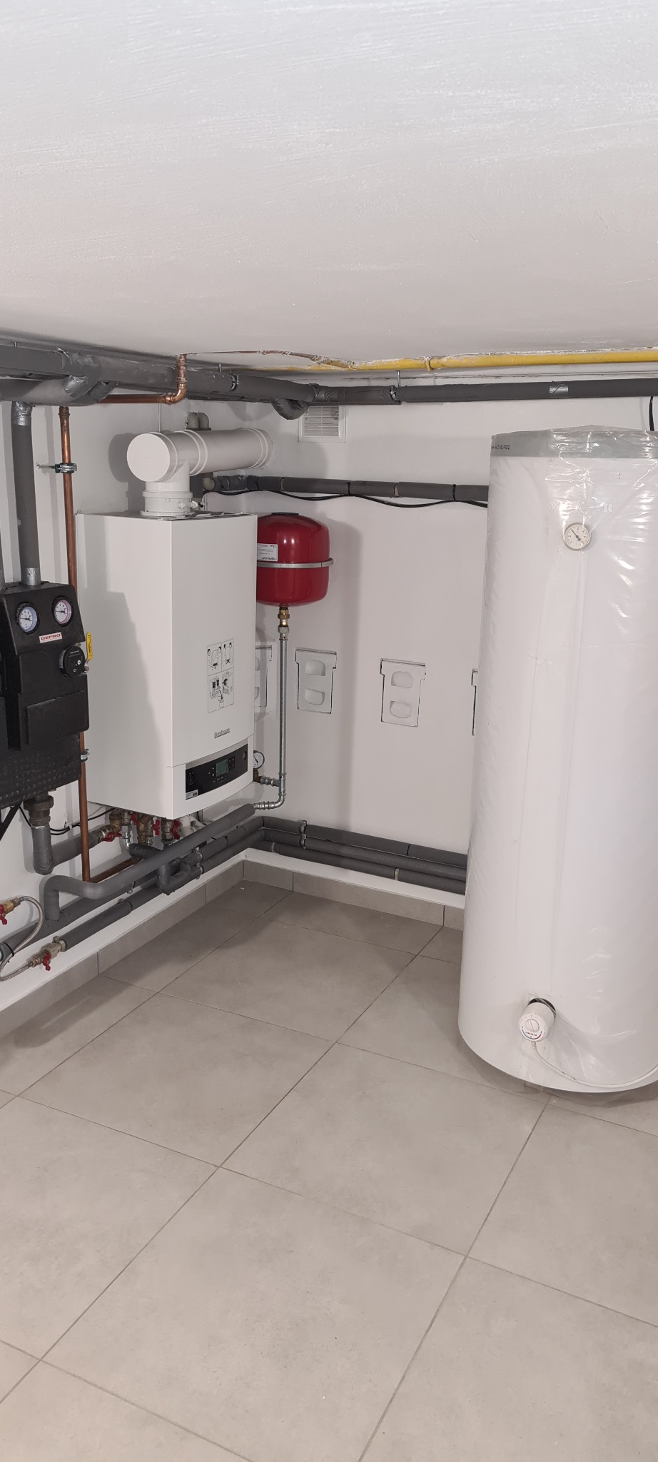 Nowoczesna kotłownia z białym kotłem gazowym Buderus, czerwonym naczyniem przeponowym i pionowym zasobnikiem ciepłej wody w folii ochronnej, widoczne rury instalacji grzewczej i czarny panel...