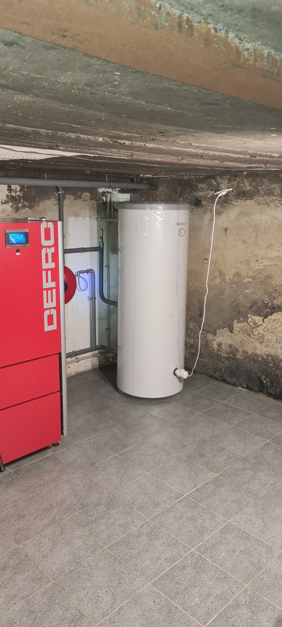 Nowy, biały zasobnik ciepłej wody, jeszcze w folii ochronnej, ustawiony obok czerwonego pieca z wyświetlaczem w surowym pomieszczeniu z szarą podłogą z płytek i nierównym sufitem.