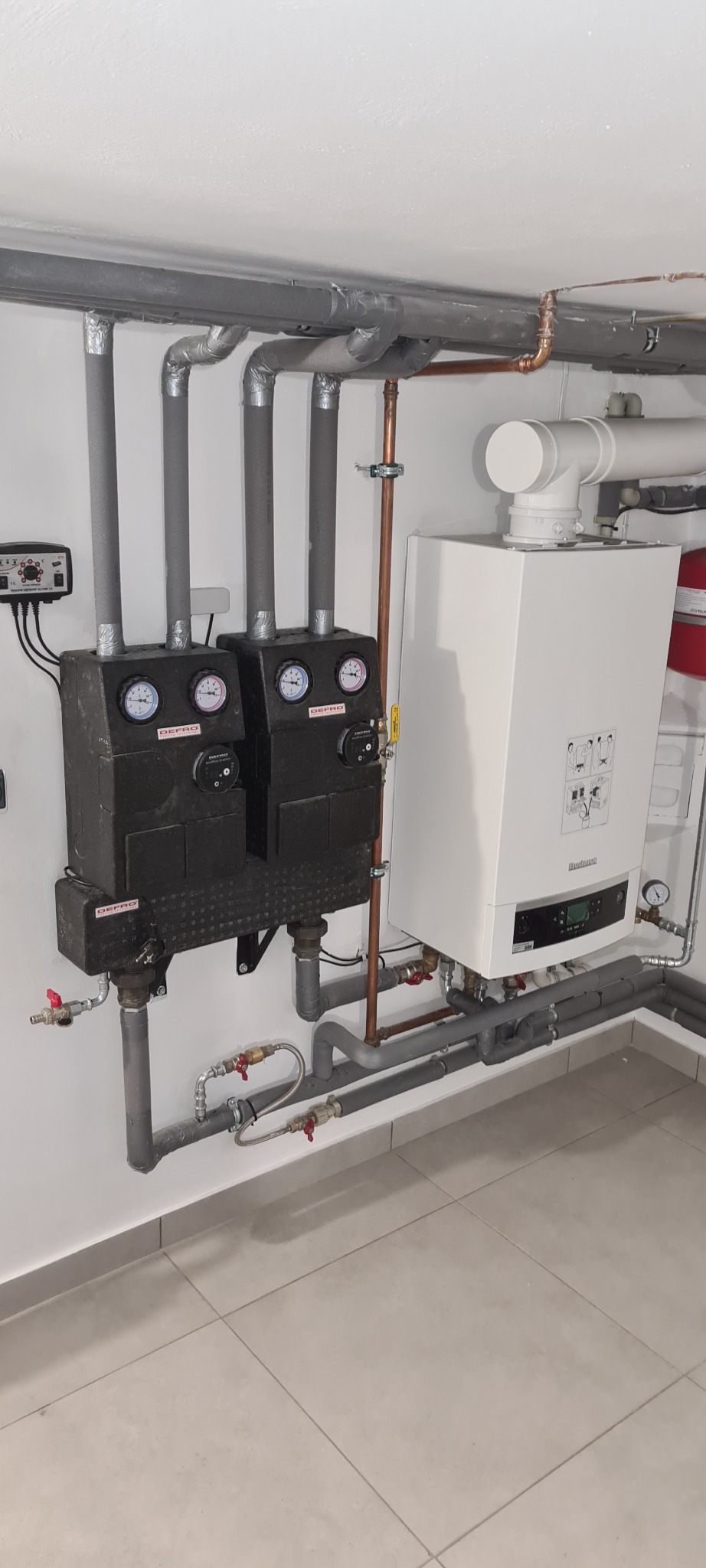 Szare rury z izolacją termiczną, czarne regulatory DEPRO z manometrami i biały wiszący piec grzewczy Buderus w kotłowni z jasnymi ścianami i podłogą.