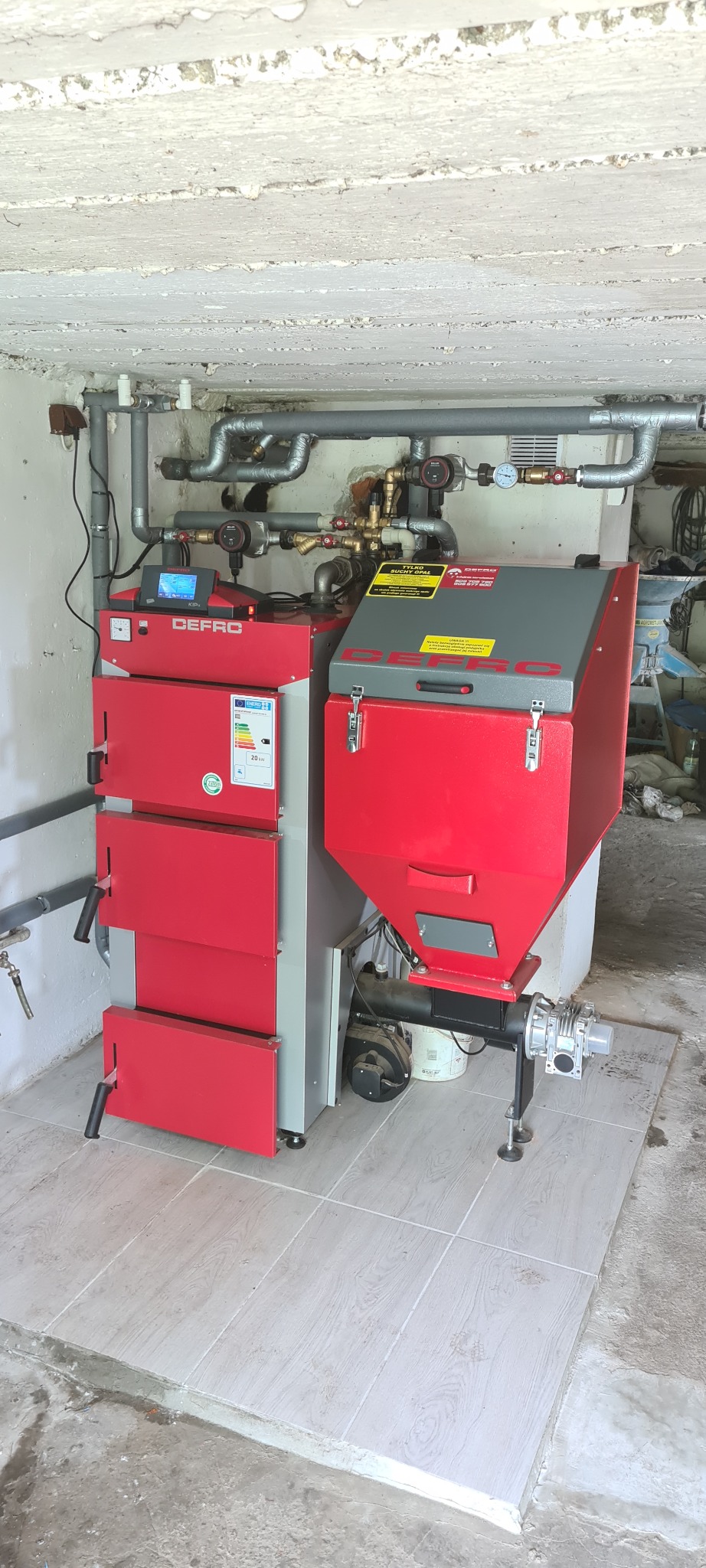 Czerwono-szary piec na paliwo stałe DEFRO z podajnikiem, zainstalowany w kotłowni z widocznymi rurami i elementami instalacji grzewczej przy suficie, na podłodze płytki imitujące drewno.