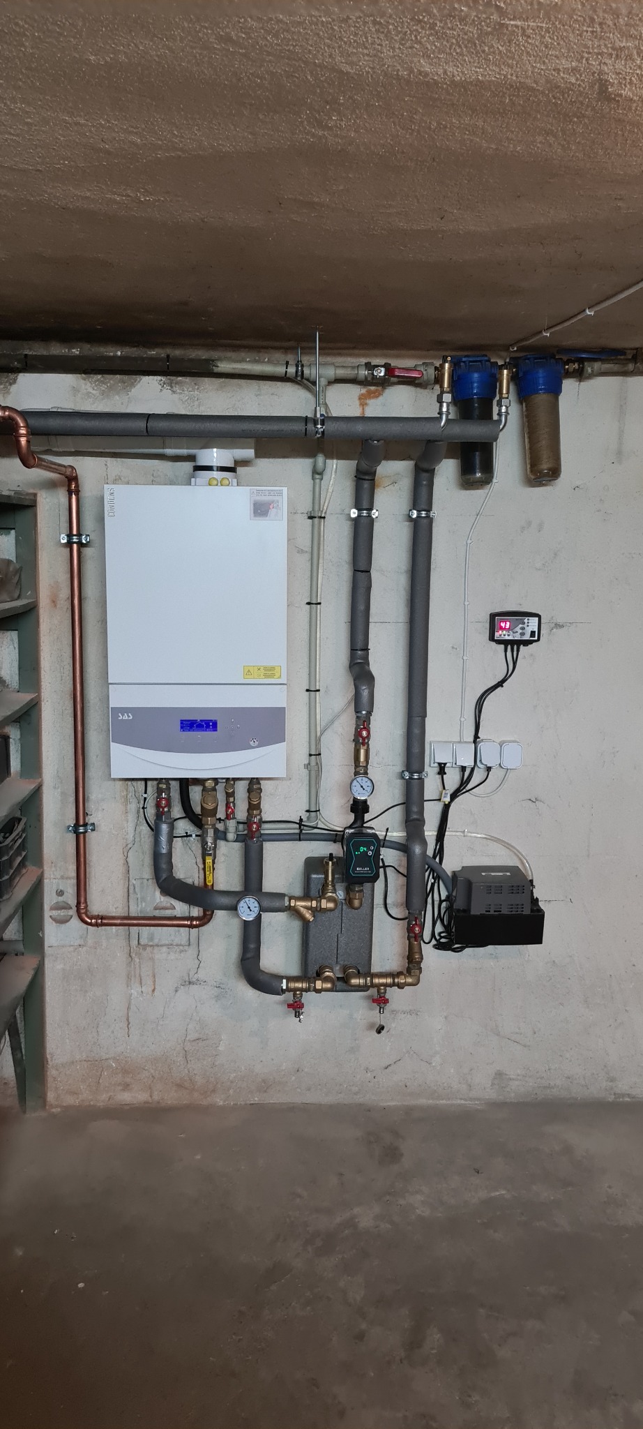 Biały piec CO SAS z wyświetlaczem cyfrowym i podłączonymi rurami miedzianymi oraz szarymi izolowanymi, zamontowany na szarej, surowej ścianie w piwnicy.