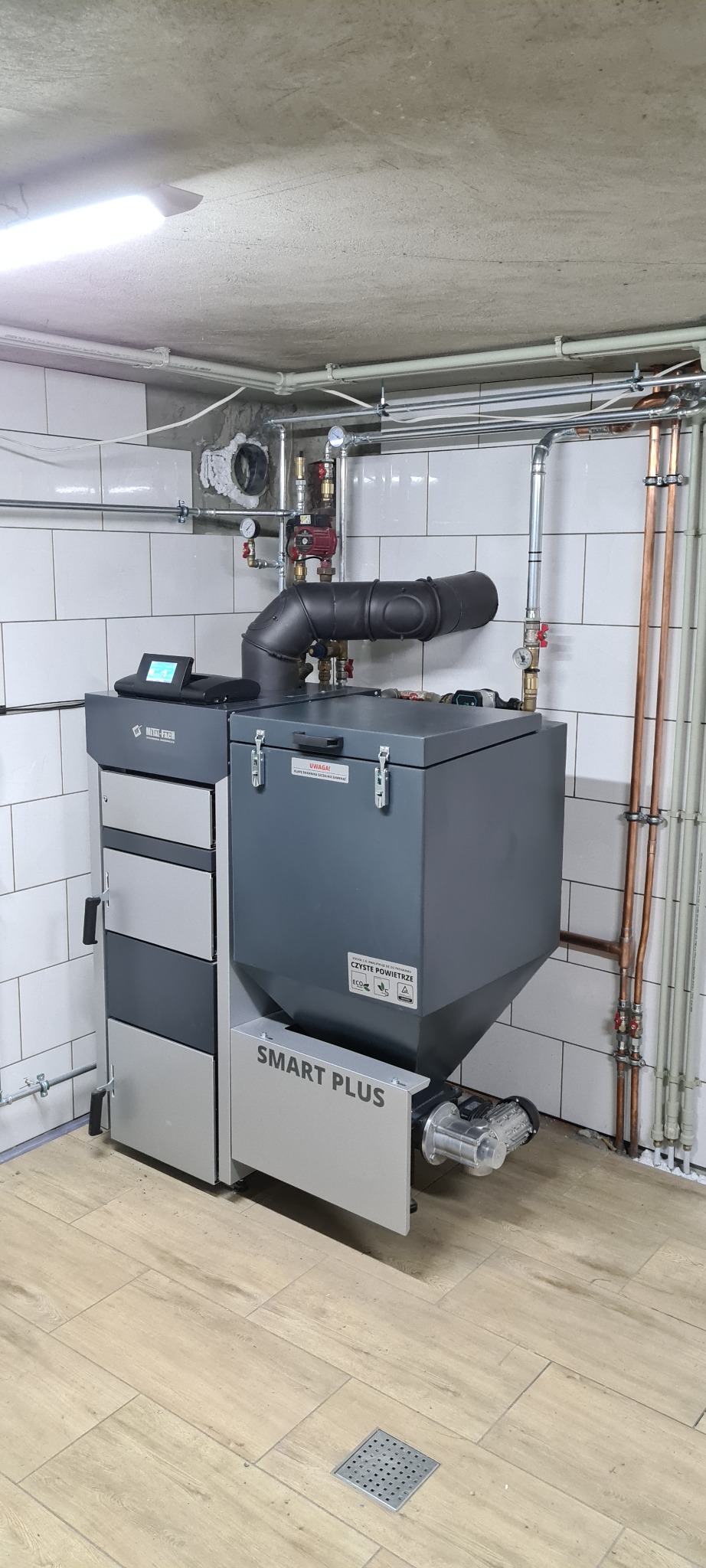 Szary piec na paliwo stałe z podajnikiem, marki Metal-Fach Smart Plus, zainstalowany w kotłowni z białymi płytkami i drewnopodobną podłogą, widoczne rury instalacji grzewczej.