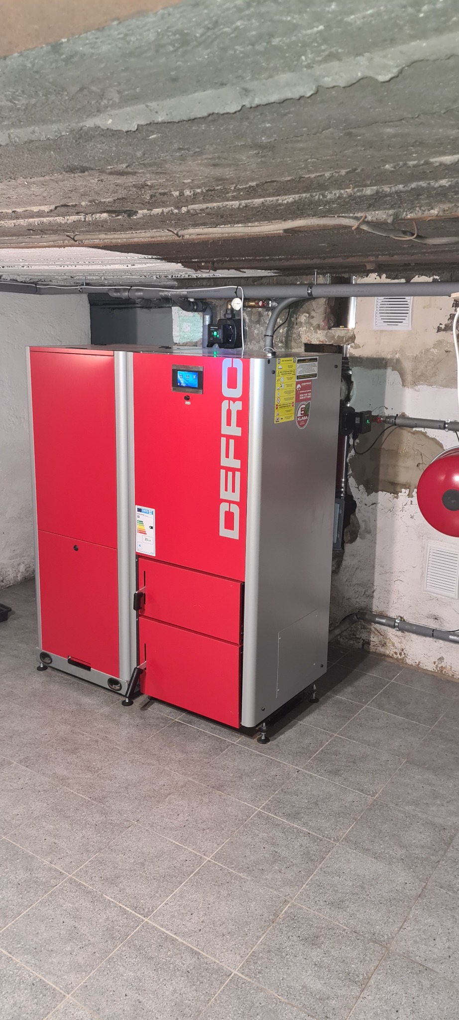 Czerwono-szary piec marki DEFRO w kotłowni z widoczną instalacją hydrauliczną i betonowym sufitem.