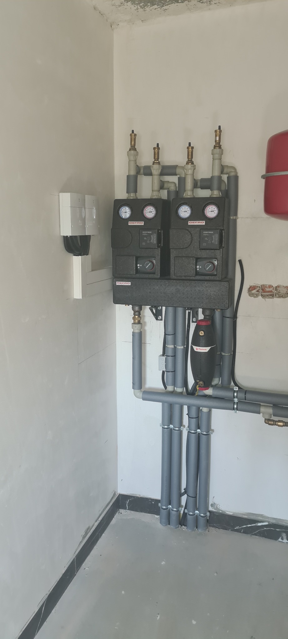 Dwa moduły sterujące ogrzewaniem DEPRO z manometrami, połączone rurami z szarą izolacją, zamontowane na ścianie w surowym pomieszczeniu z widocznymi śladami wilgoci na suficie.