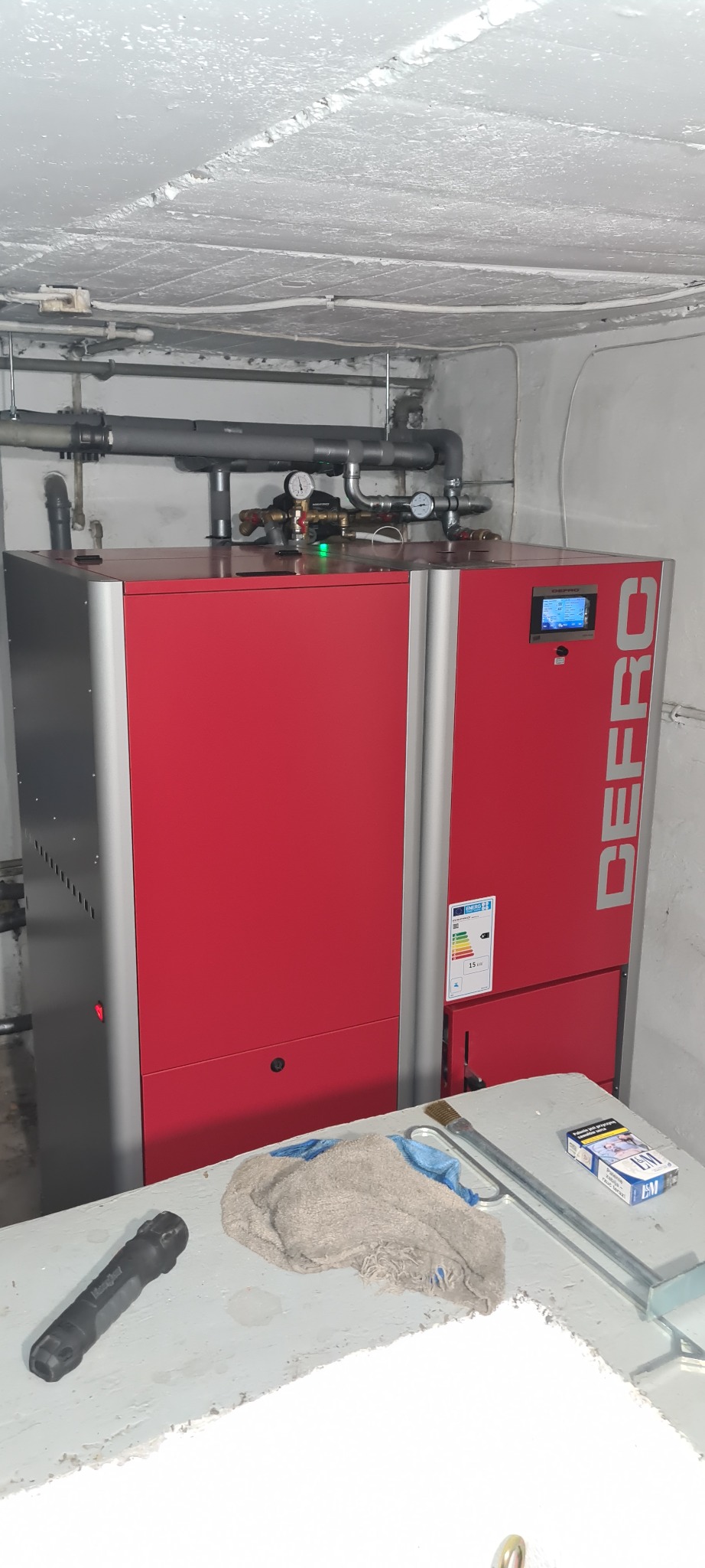 Czerwony piec z szarymi elementami w kotłowni, widoczne rury, manometry, sterownik, etykieta energetyczna, na blacie leżą narzędzia i szmatka.
