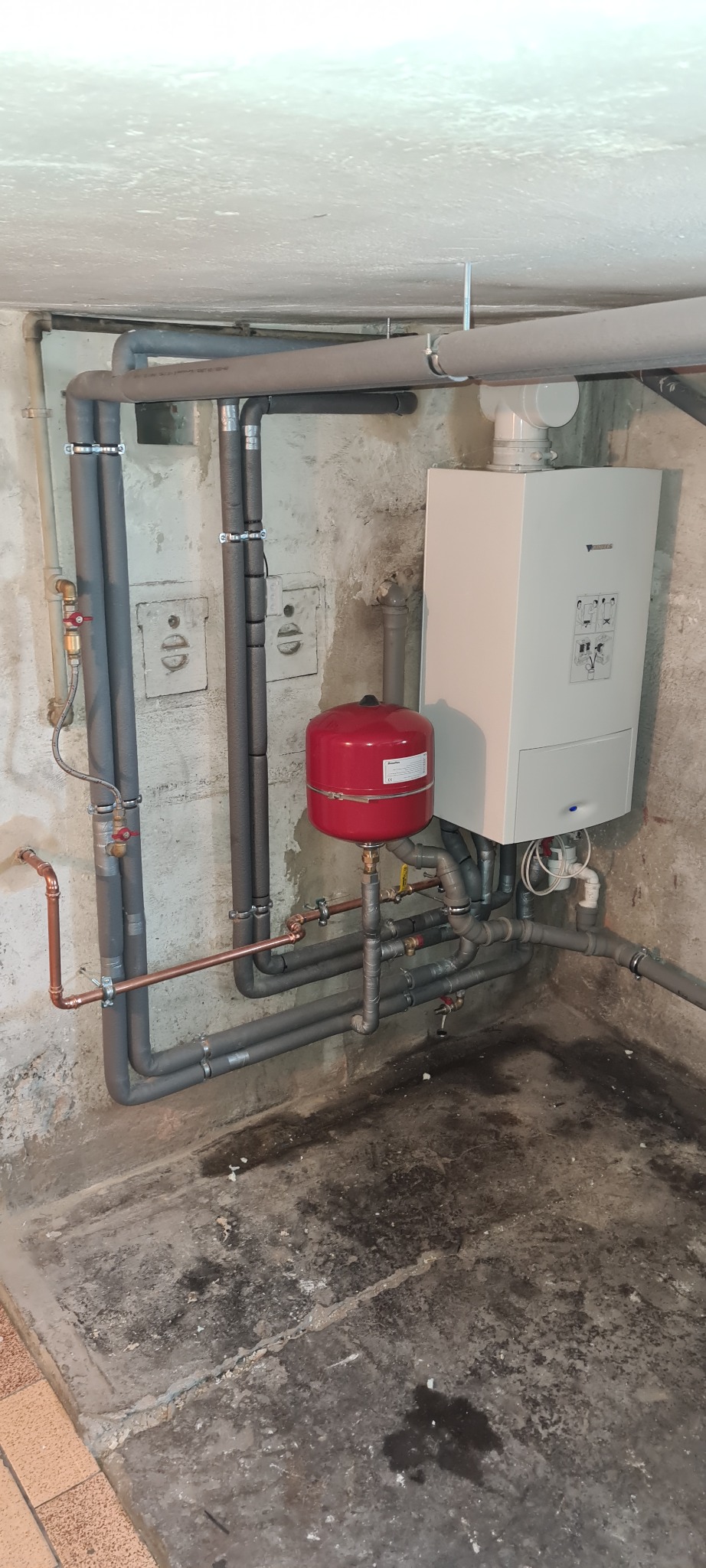 Instalacja grzewcza w piwnicy: biały piec gazowy Junkers, czerwony zbiornik wyrównawczy i szare rury z izolacją, zamontowane na surowej ścianie i podłodze.