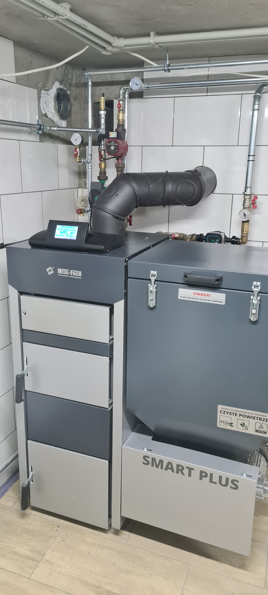 Szary piec na paliwo stałe marki Metal-Fach Smart Plus z czarnym kominem i widoczną instalacją hydrauliczną na tle białych kafelków.