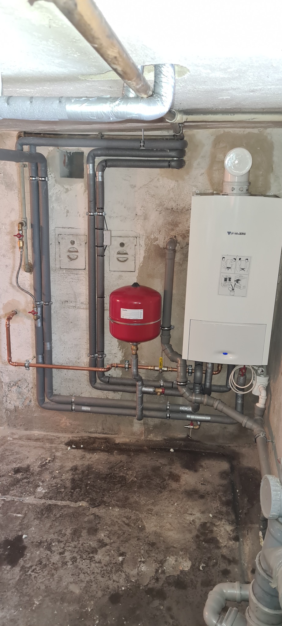 Zmontowany, wiszący piec gazowy Junkers z czerwonym zbiornikiem wyrównawczym, licznymi rurami miedzianymi i szarymi rurami PCV na tle surowej ściany i posadzki piwnicy.