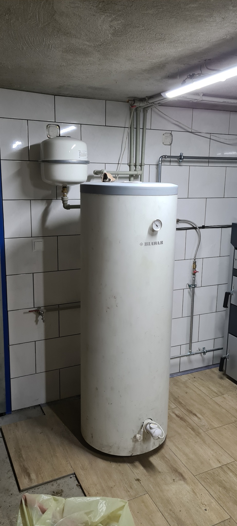 Biały, cylindryczny zasobnik ciepłej wody z manometrem, zamontowany w pomieszczeniu z białymi płytkami i widoczną instalacją hydrauliczną.