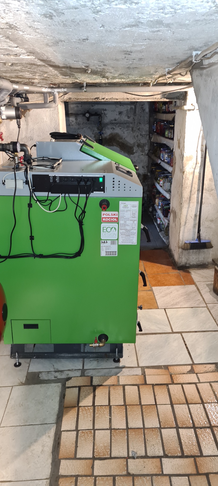 Zielony piec na paliwo stałe SAS w piwnicy z instalacją grzewczą i regałem z przetworami, widoczne tabliczki znamionowe i certyfikaty.