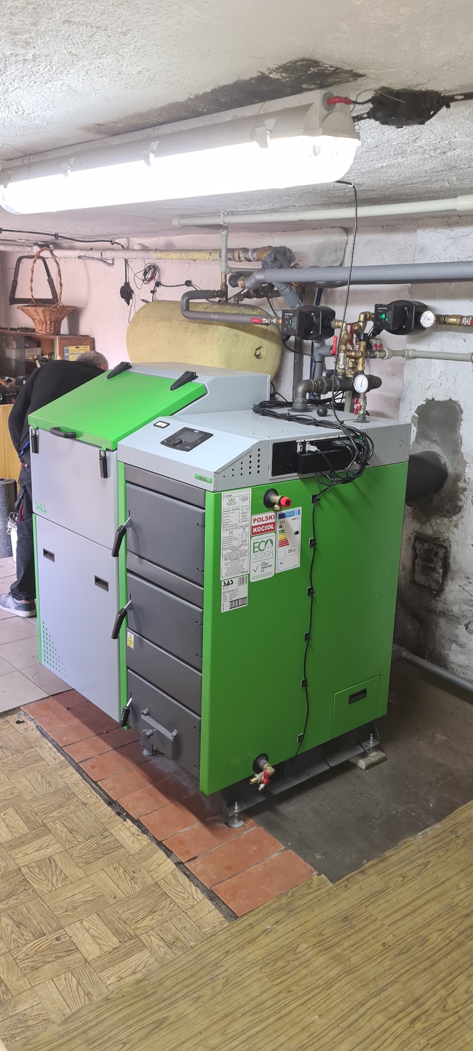 Szaro-zielony piec na paliwo stałe marki SAS, z otwartą górną klapą, zainstalowany w piwnicy z widocznymi rurami i armaturą grzewczą, na podłodze z terakoty i parkietu.