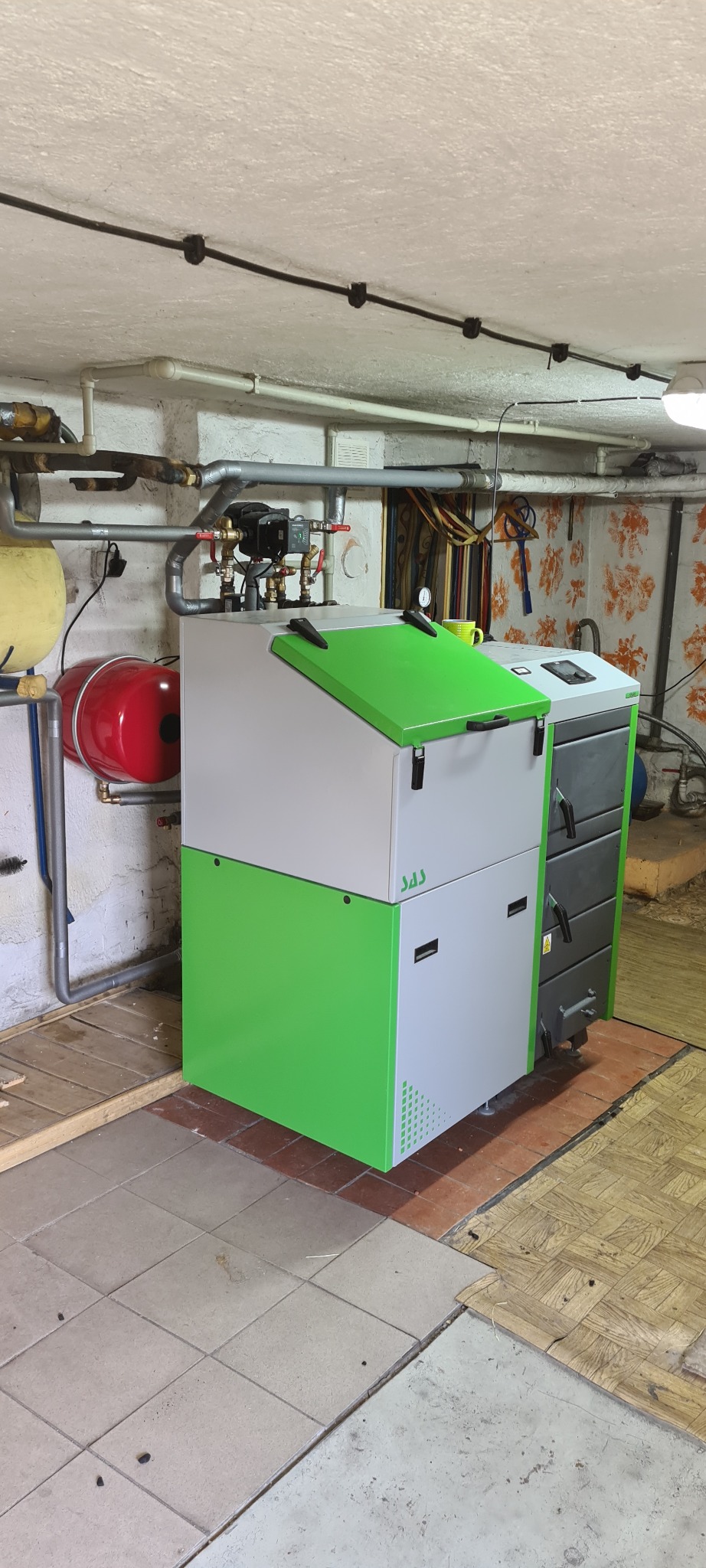 Nowoczesny piec na paliwo stałe SAS w kotłowni, z widocznymi rurami instalacji grzewczej i zbiornikiem wyrównawczym, na podłodze zróżnicowane płytki i drewno.