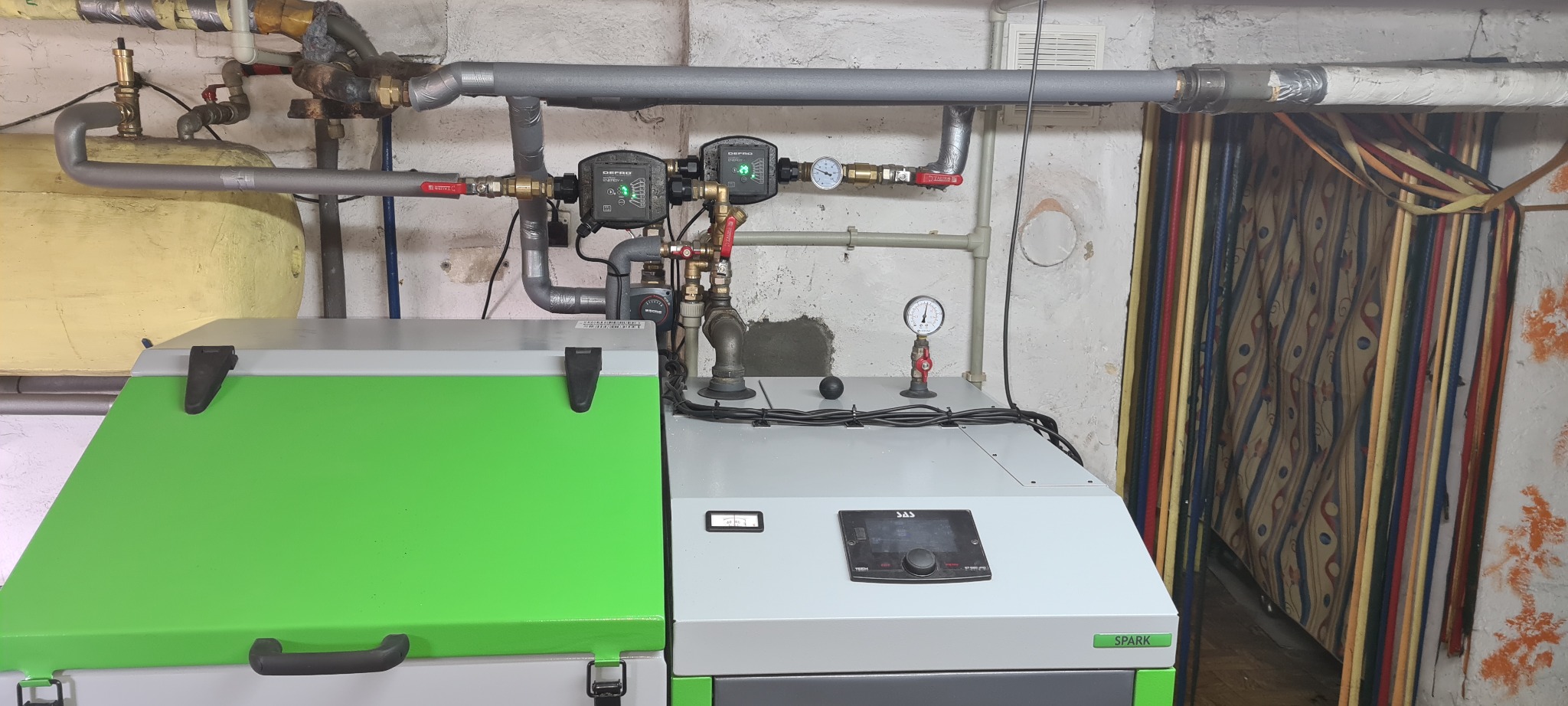 Nowoczesny piec na paliwo stałe z zielonymi akcentami, podłączony do instalacji CO z widocznymi zaworami i manometrami, umieszczony w pomieszczeniu z surowymi ścianami.