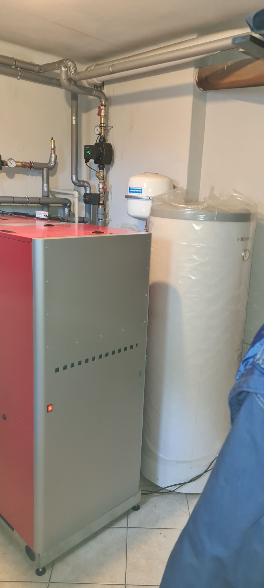 Kotłownia z piecem na paliwo stałe z podajnikiem, obok biały zasobnik wody owinięty folią ochronną, widoczne rury instalacji grzewczej pod sufitem.