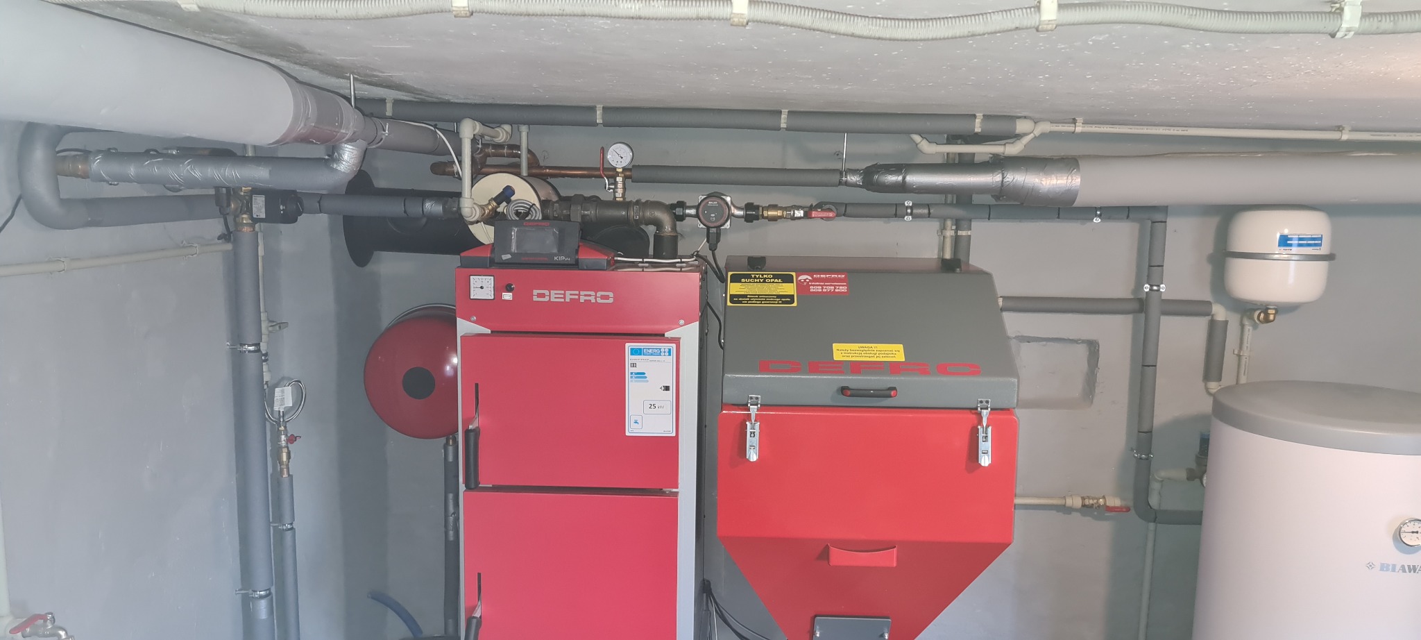 Zainstalowany kocioł na paliwo stałe marki Defro w kotłowni, z widocznymi rurami instalacji grzewczej i zbiornikiem wyrównawczym.