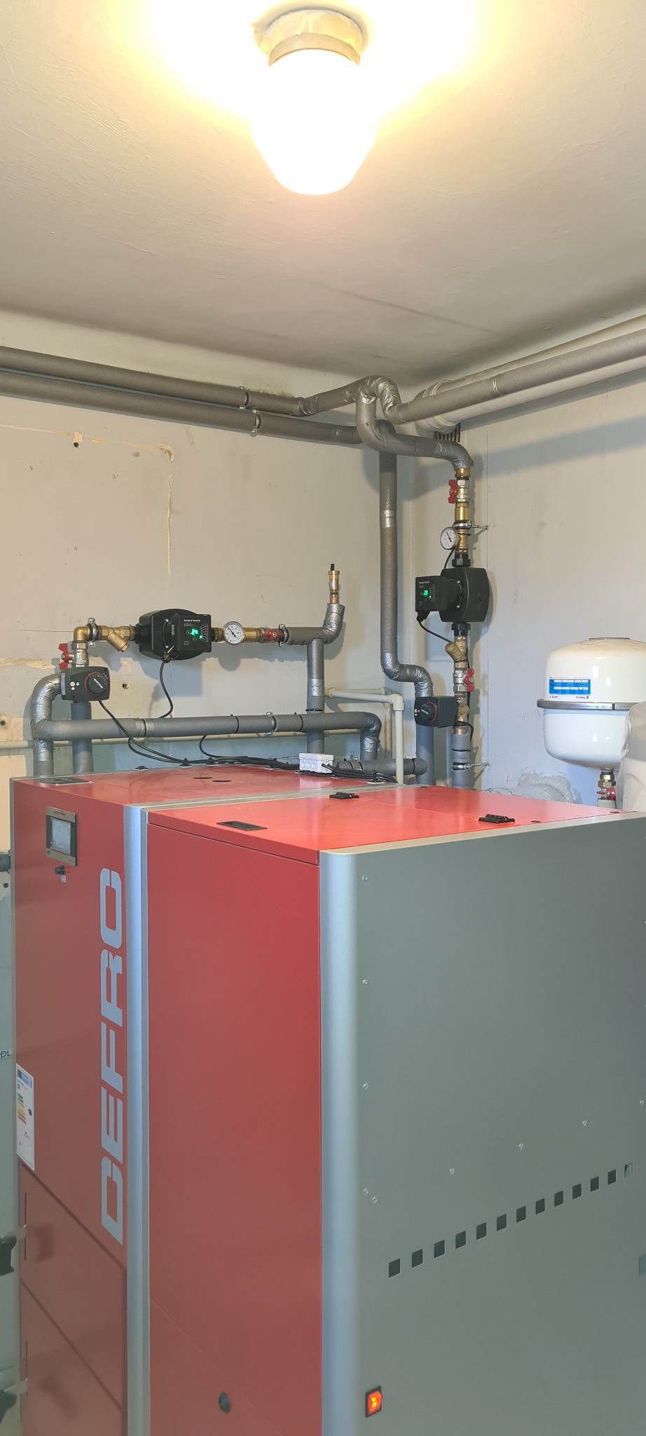 Zainstalowany, nowoczesny piec DEFRO z kompletną instalacją hydrauliczną, w tym pompy obiegowe i orurowanie, zamontowany w pomieszczeniu kotłowni z widocznym oświetleniem sufitowym.