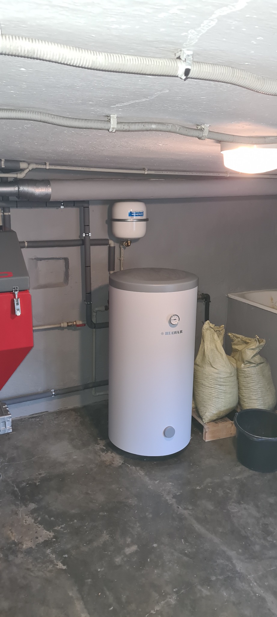 Biały, cylindryczny zasobnik ciepłej wody użytkowej Biawar z manometrem, zamontowany w kotłowni z workami i elementami instalacji grzewczej.