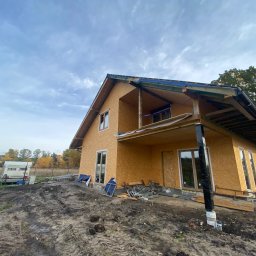 ND-BUD Nazarii Doroshchuk - Dwukondygnacyjny dom w trakcie budowy z płyt OSB, widoczne elementy konstrukcyjne, okna i teren budowy z przyczepą kempingową w tle.