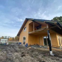 Dwukondygnacyjny dom w trakcie budowy z płyt OSB, widoczne elementy konstrukcyjne, okna i teren budowy z przyczepą kempingową w tle.