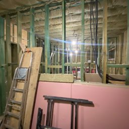 ND-BUD Nazarii Doroshchuk - Wnętrze domu szkieletowego w trakcie budowy z widoczną izolacją stropu, ścianami z drewnianej ramy, instalacją elektryczną, opartą drabiną i rusztowaniem.