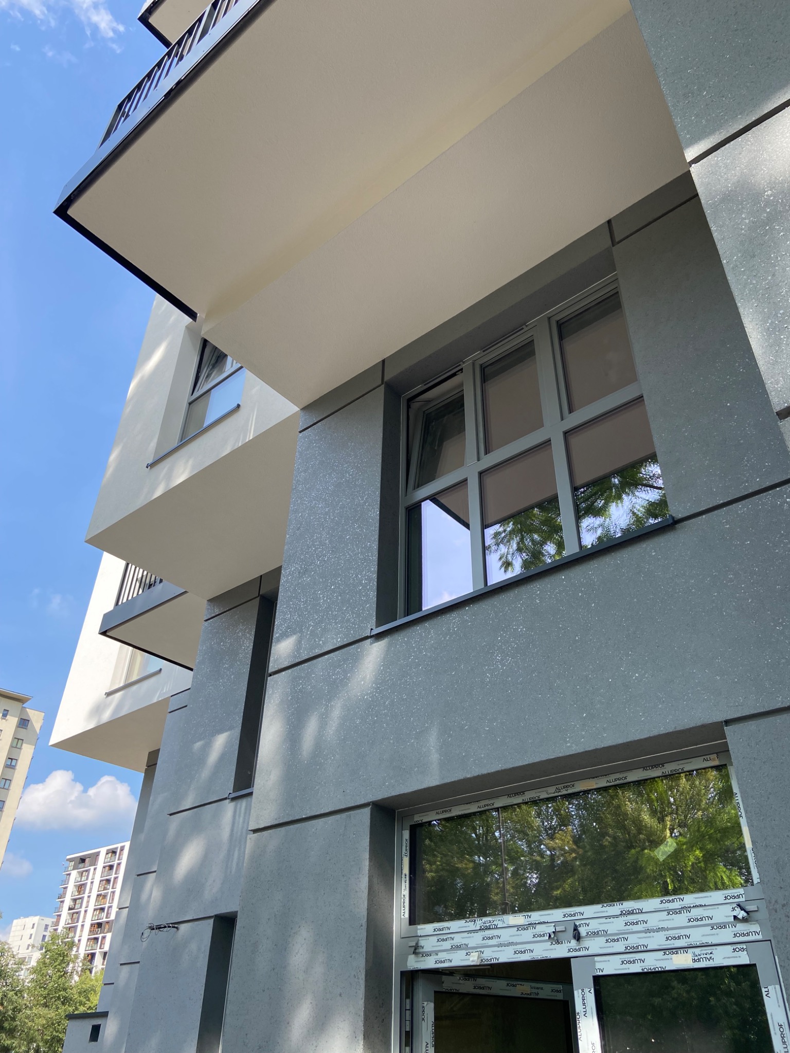 Fragment nowoczesnego budynku wielorodzinnego z elewacją w odcieniach szarości i bieli, widok z dołu, z oknami z ramami Alufrof i odbiciem drzew w szybach.
