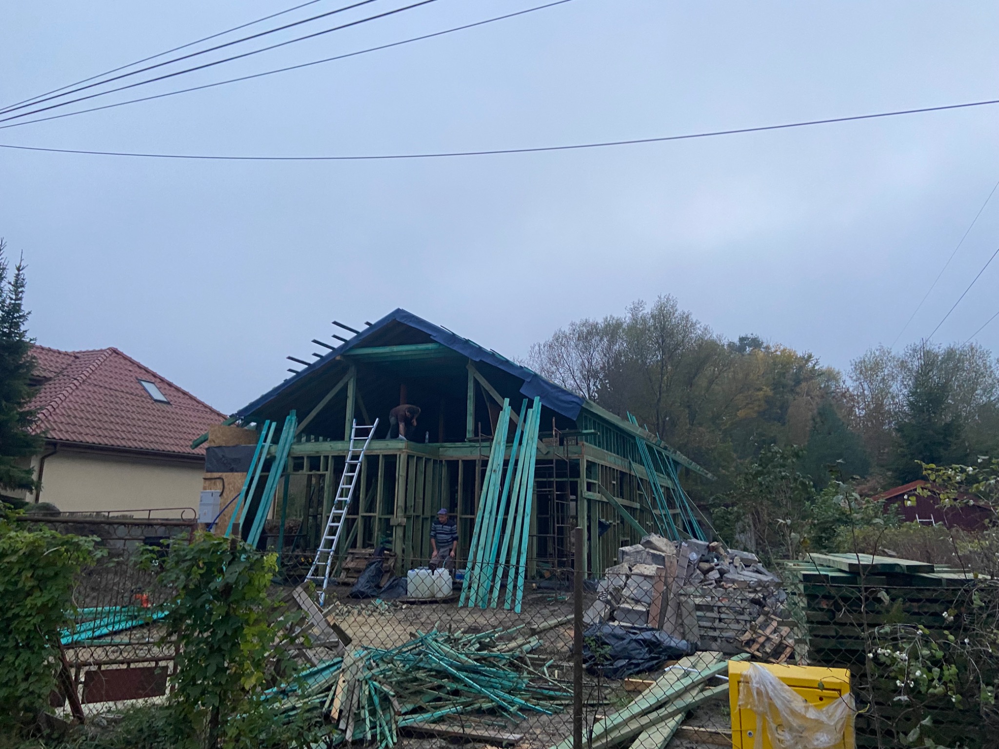 Konstrukcja szkieletowa domu w trakcie budowy, widoczne pionowe i poziome elementy drewniane, drabina oparta o ścianę, dwóch pracowników na rusztowaniu, materiały budowlane i narzędzia na ziemi.